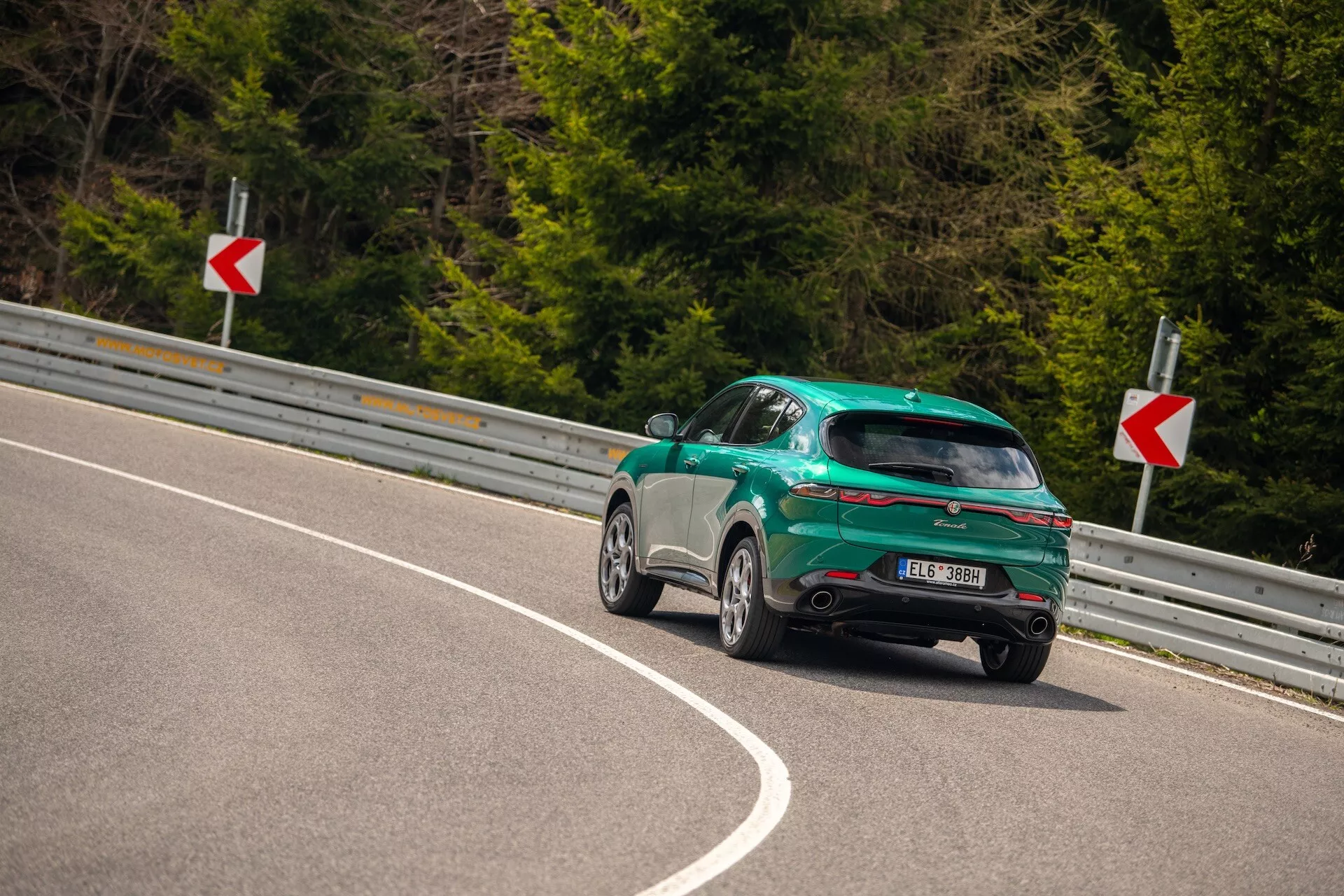 Alfa Romeo Tonale 2022 zachycené z úhlu tří čtvrtin zezadu na zakřivené silnici, zdůrazňující sportovní design s karoserií hatchback, lesklý finiš a aerodynamické prvky včetně zadního spoileru, slitinových kol a LED zadních světel.