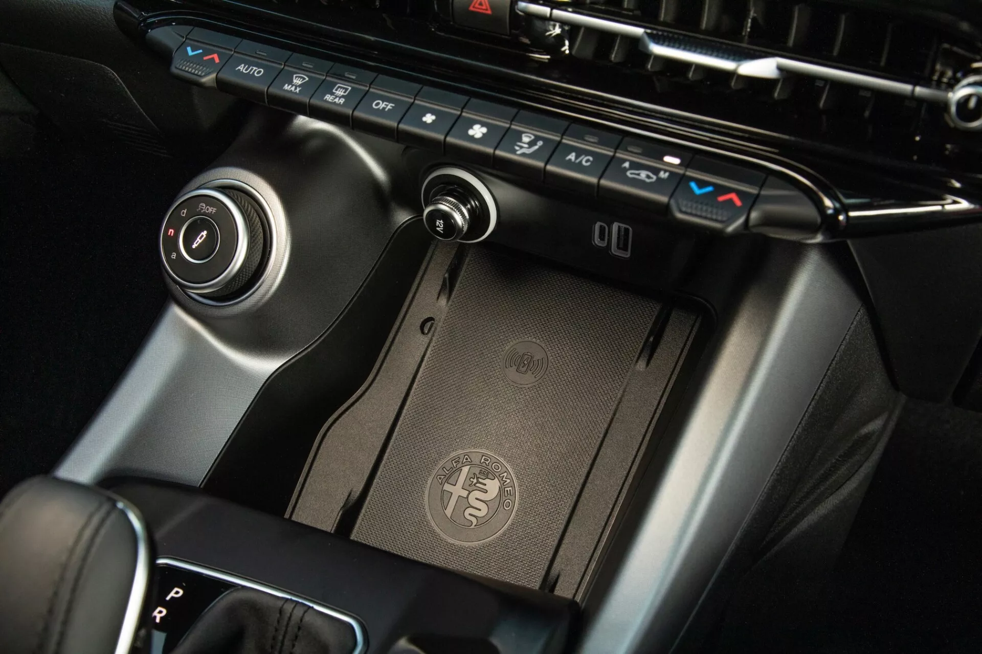 Detailní pohled na středovou konzoli Alfa Romeo Tonale 2022, zobrazující panel klimatizace, bezdrátovou nabíjecí podložku, volič převodů a ovládací kotouč infotainmentu s prémiovým provedením a moderním designem.