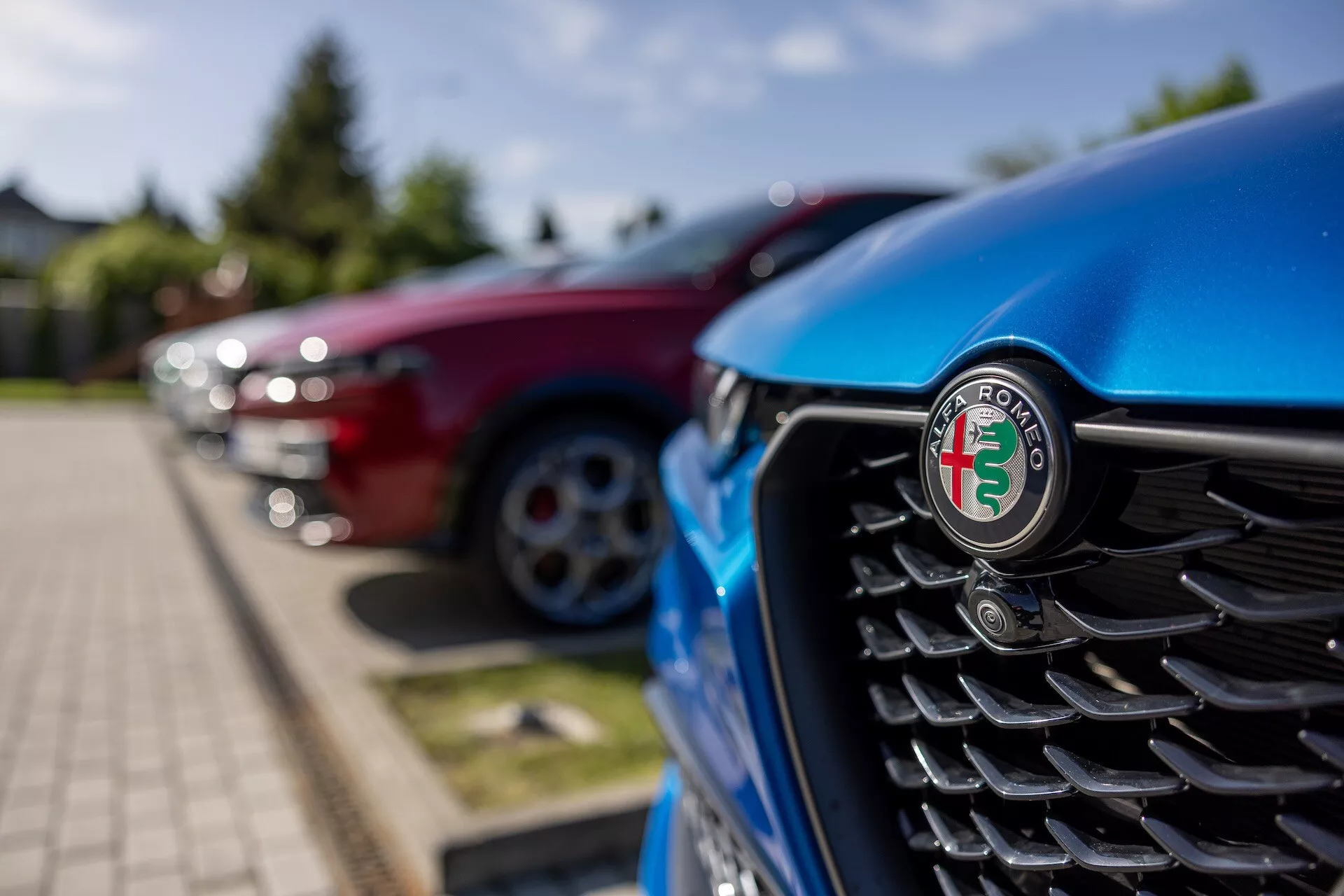 Detailní pohled na přední mřížku modrého Alfa Romeo Tonale 2022 se zaměřením na emblem, který obsahuje vertikální lamely a rozmazané pozadí s dalšími vozidly