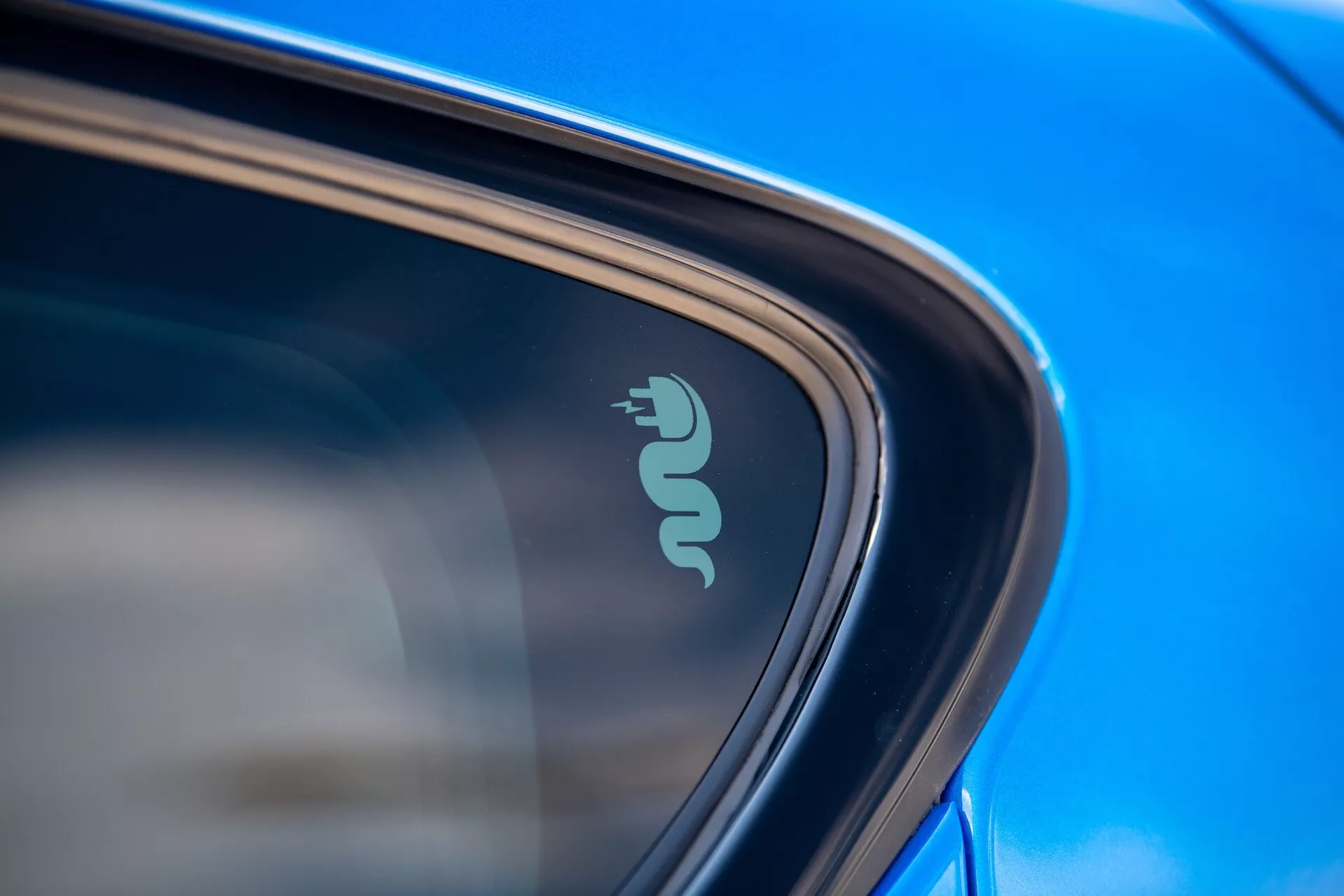 Detailní záběr na technologický odznak Alfa Romeo Tonale 2022 naznačující elektrickou nebo hybridní schopnost poblíž okna auta, zdůrazněný odrazivým modrým lakem a elegantním designem na přirozeném denním světle