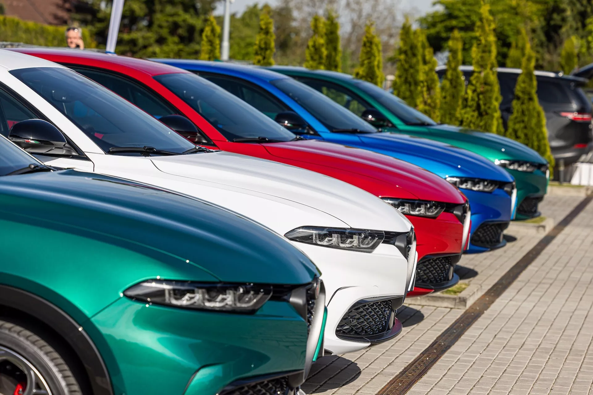 Řada vozidel Alfa Romeo Tonale 2022 zaparkovaná diagonálně s výhledem z předního tříčtvrtečního úhlu zdůrazňující LED světlomety, různé designy mřížek, lesklé lakování a slitinová kola na pozadí jasného slunečního svitu a zeleně