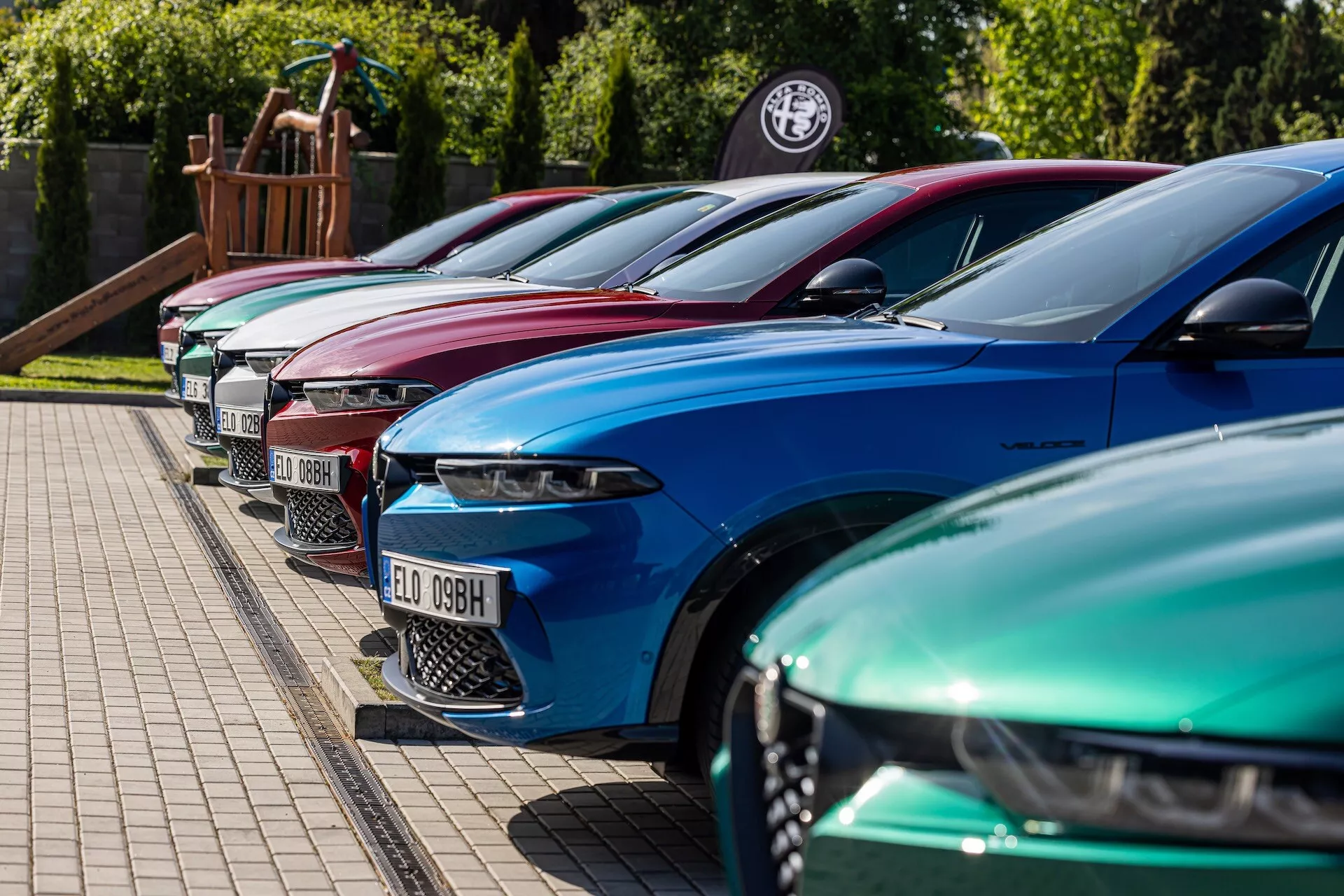 Řada Alfa-Romeo Tonale z roku 2022 ve výrazných barvách ukazuje luxusní design s pokročilými LED světlomety, zaparkované v diagonální formaci s důrazem na přední levou stranu z nízkého až středního úhlu na zpevněné ploše vedle travnatého okraje, možná u dealera nebo na akci nadšenců automobilů