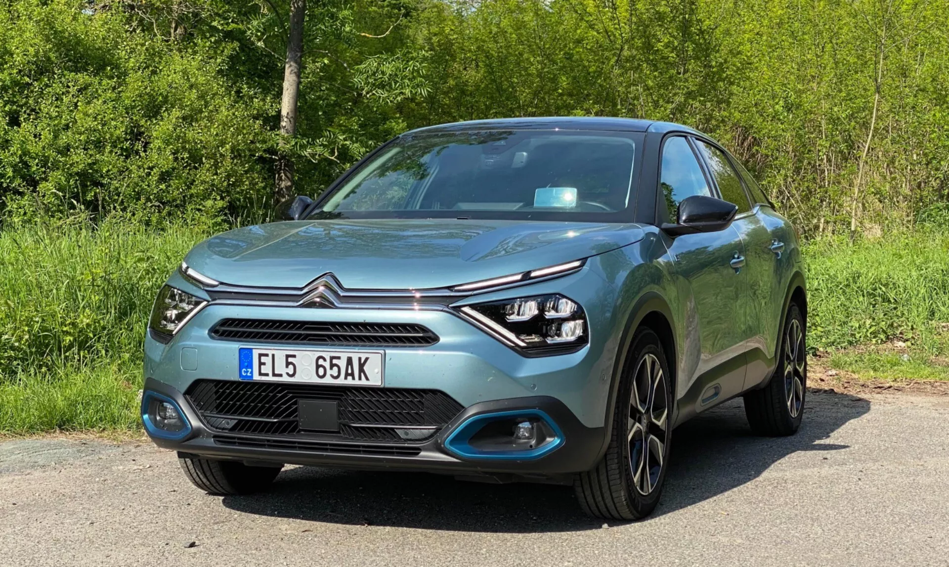 Obrázek moderního hatchbacku Citroen C4 (E-C4) z roku 2020 z nízkoúhlového předního tříčtvrtečního pohledu, ukazuje přední část, řidičovu stranu, kapotu, světlomety a design kol. Vybavení zahrnuje LED denní svícení kolem elegantních světlometů, mlhovky integrované do nárazníku, centrální mřížku s logem výrobce, lemované chromovými ozdobami, hladké linie kapoty, kliky dveří a zrcátka sladěné s barvou karoserie, vícepaprsková litá kola, metalický lak a přední registrační značku. Auto je zaparkované u silnice s zeleným porostem v pozadí.