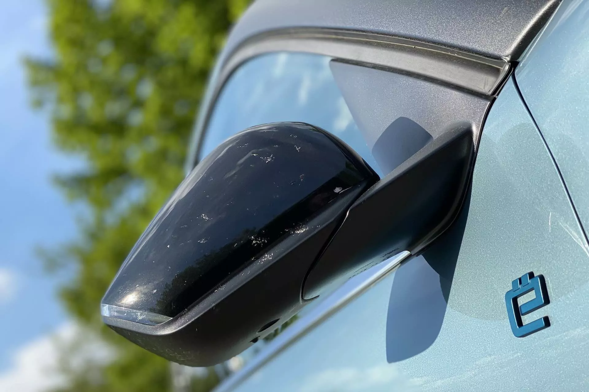 Detailní pohled na zpětné zrcátko na Citroenu C4 2020 (E-C4), zdůrazňující černé pouzdro zrcátka a kontrastující světlou barvu karoserie. V pozadí je vidět jasná obloha a zeleň, což naznačuje venkovní prostředí. Stav auta je nový, ačkoli na pouzdru zrcátka je vidět nějaká špína. Viditelný je také stylizovaný emblém 'e', který možná naznačuje elektrický model.