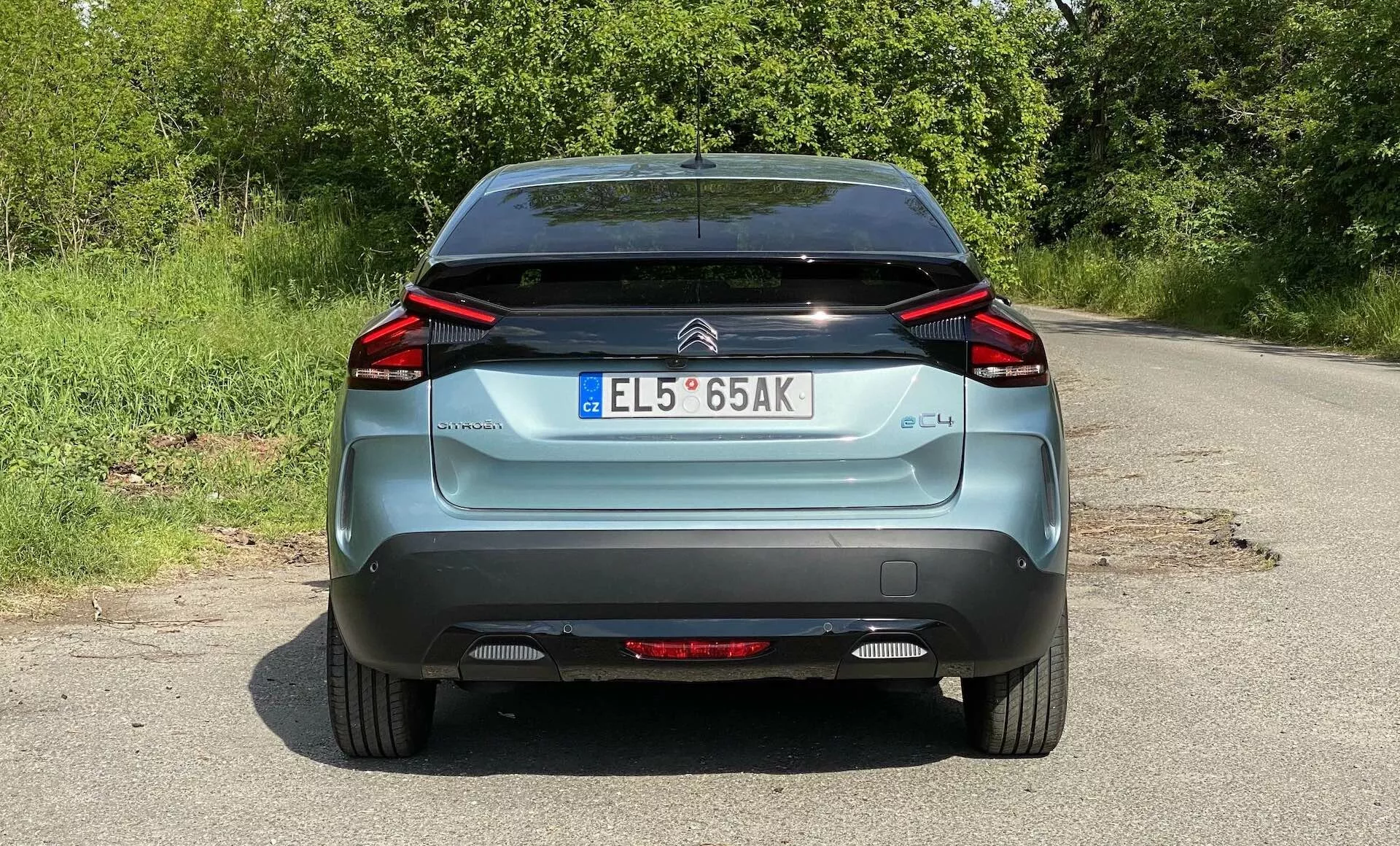 Pohled zezadu na modrý Citroen C4 z roku 2020 (e-C4), zaparkovaný na venkovské silnici. Obrázek zachycuje celou šířku vozidla od spodku kol po střechu, ukazuje výrazná úhlová LED zadní světla, znak značky, označení 'C4', evropskou registrační značku, stěrač zadního okna a černou spodní část nárazníku. Viditelné jsou také odrazky a zadní mlhové světlo.