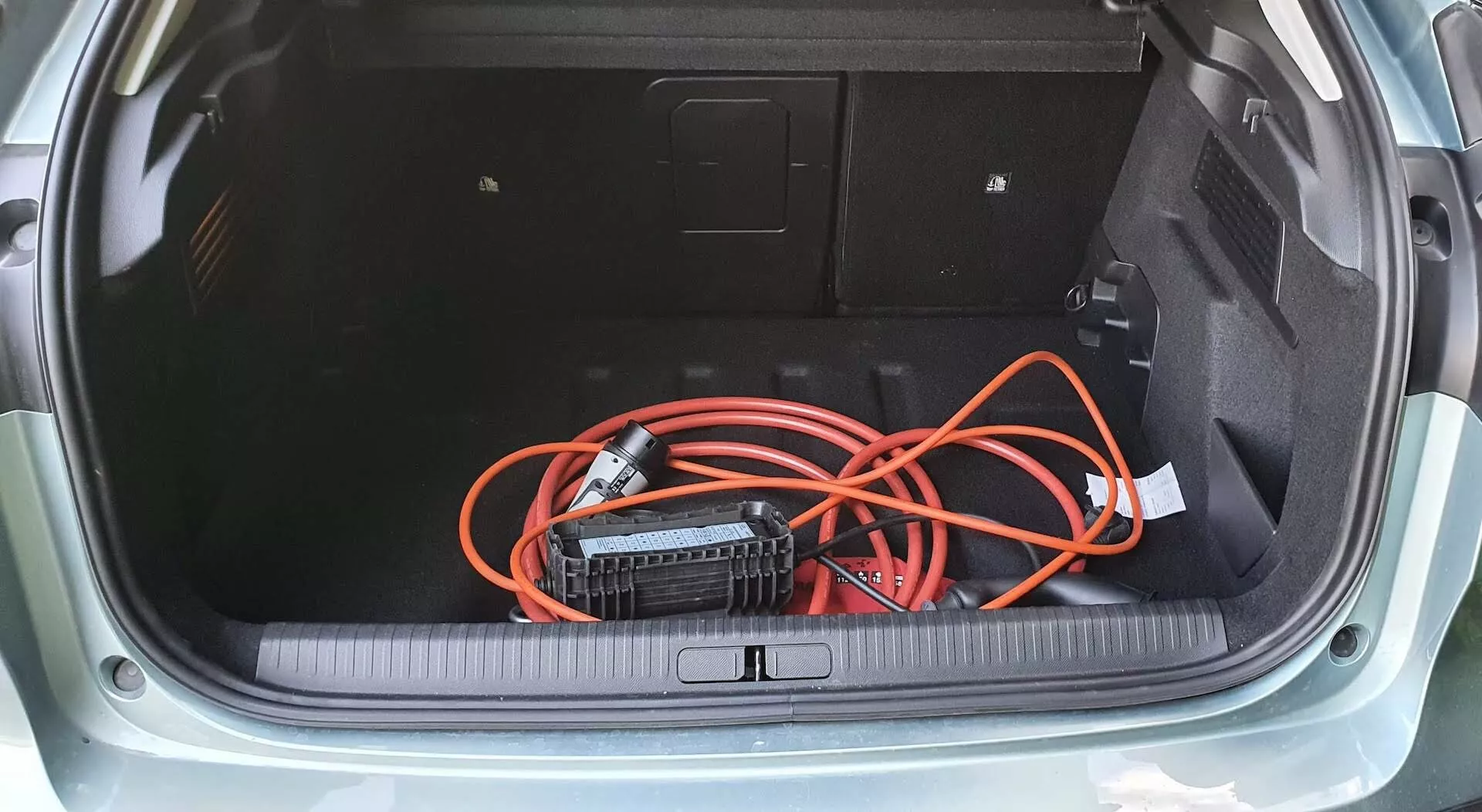 Pohled shora do zavazadlového prostoru hatchbacku Citroen C4 2020 (e-C4) s kobercovým dnem, bočními panely s přístupovým panelem a volně vloženým oranžovým prodlužovacím kabelem připojeným k přenosnému napájecímu měniči. Prahová hrana kufru má ochranný kryt z gumy nebo plastu, což zdůrazňuje praktický design úložného prostoru vozu.