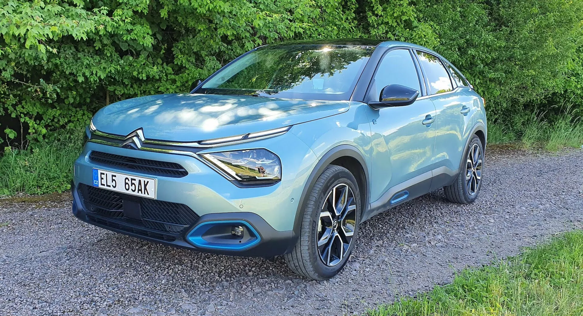 Světlé modrý Citroen C4 2020 (e-C4) kompaktní SUV zaparkované na štěrku s vegetací v pozadí, přední čtvrtinový pohled zobrazující stranu řidiče, přední část, tvarovanou karoserii, LED světlomety, vícepaprsková litá kola, přední mřížku a odznak výrobce, zdůrazňující designové a technologické prvky.