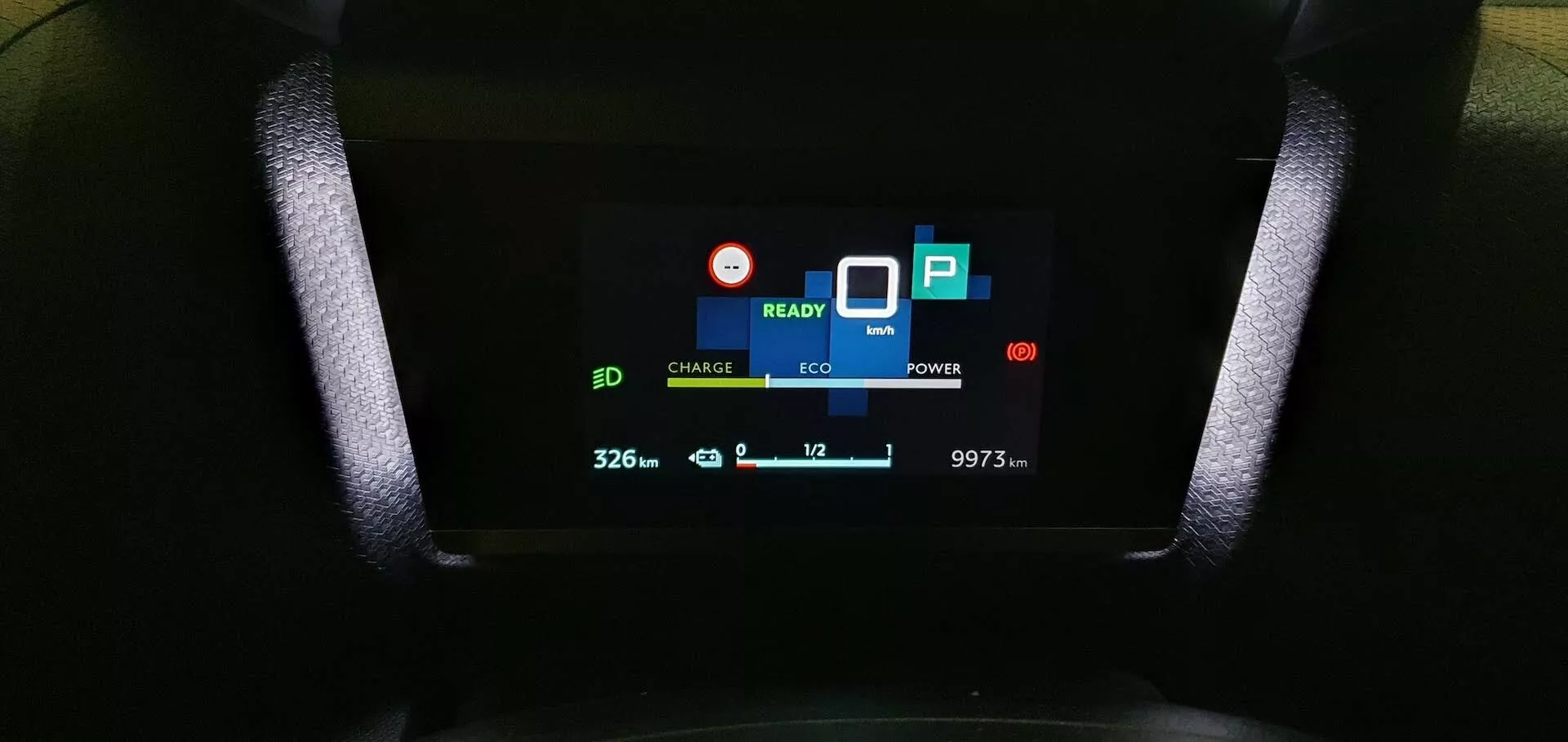 Blízký pohled na digitální přístrojový panel Citroen C4 2020 (e-C4) zobrazující indikátory 'CHARGE' a 'ECO', signál 'READY', ikonu baterie s hodnotou '32.6', ikonu listu označující ekologický režim, indikátor spotřeby energie 'POWER', symbol parkování 'P', '10' se šipkou dolů indikující teplotu a stav kilometrů '9973 km', zachycený z pohledu řidiče.