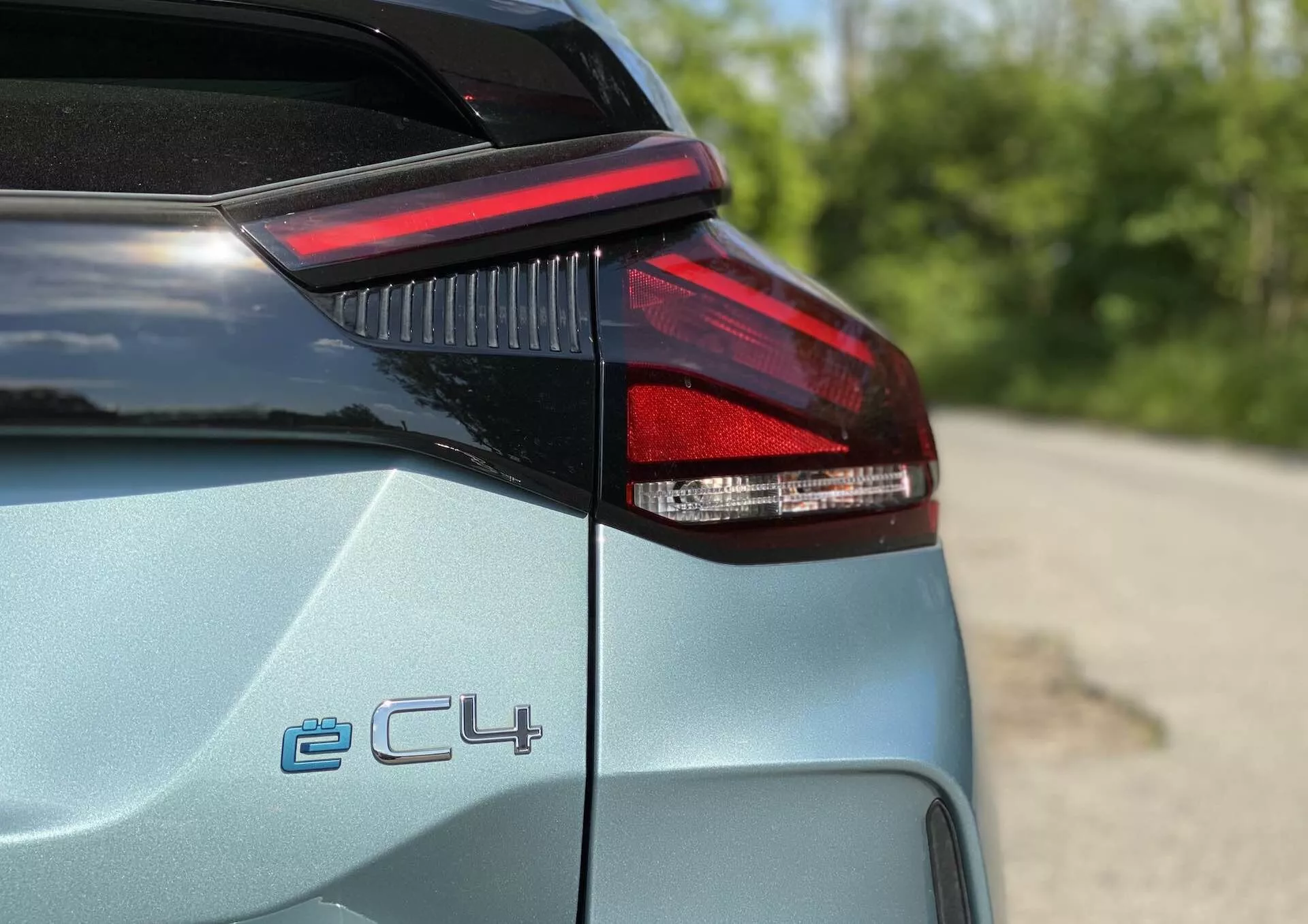 Detailní pohled na zadní levou stranu Citroen C4 2020 (e-C4), zobrazující LED zadní světlo, okraj kufru a část zadního panelu; značka 'ë-C4' označující elektrickou variantu; světle kovová barva s lesklým povrchem; mělká hloubka ostrosti, rozmazané venkovní pozadí se zelení.