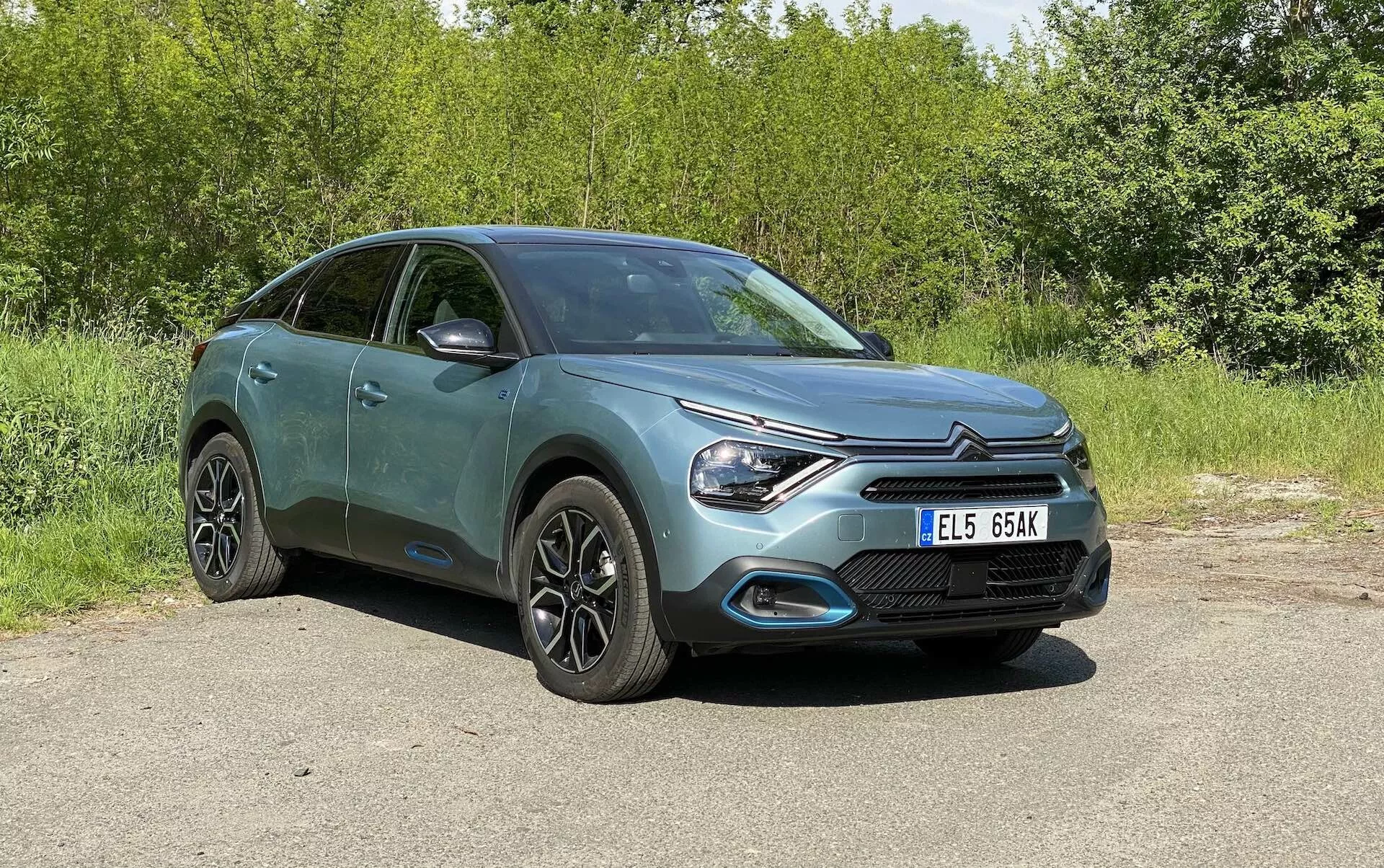 Kompletní pohled na Citroën C4 2020 (e-C4) zaparkovaný venku, pořízený z předního tříčtvrtečního úhlu, ukazující přední mřížku, elegantní LED světlomety a tvar kapoty, částečně boční profil včetně dveří a bočních zrcátek. Hatchback s crossover/SUV konstrukcí má moderní litá kola, integrované ukazatele směru v zrcátkách a metalický lak. Pozadí je zelené, auto je zobrazeno na denním světle bez rušivých prvků.