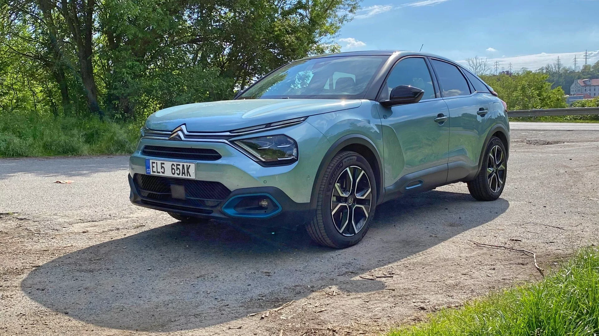 Obrázek zobrazuje hatchback nebo kompaktní SUV Citroen C4 (e-C4) z roku 2020 z nízkého předního tříčtvrtinového úhlu. Tento pohled zdůrazňuje přední část, včetně mřížky chladiče, světlometů a nárazníku, stejně jako profil na straně řidiče, který zobrazuje přední blatník, kolo, dveře řidiče a část zadních dveří. Auto má moderní, sportovní design s hladkými liniemi, šikmou střechou, výraznými LED světlomety a prominentní mřížkou chladiče. Přední nárazník má kontrastní černé lemování, zvýrazňující jeho drsný vzhled. Složitý design kol s mnoha paprsky naznačuje zaměření na styl a výkonnost. Metalická barva se na slunci jasně leskne, odrážejíc čistý a leštěný exteriér. Vozidlo je zaparkované venku v zelené krajině, jak naznačují stromy a jasné povětrnostní podmínky. Technologické prvky jako senzory nebo kamery nejsou z tohoto úhlu potvrzeny a poznávací značka je viditelná, ale pro zachování soukromí je vynechána.