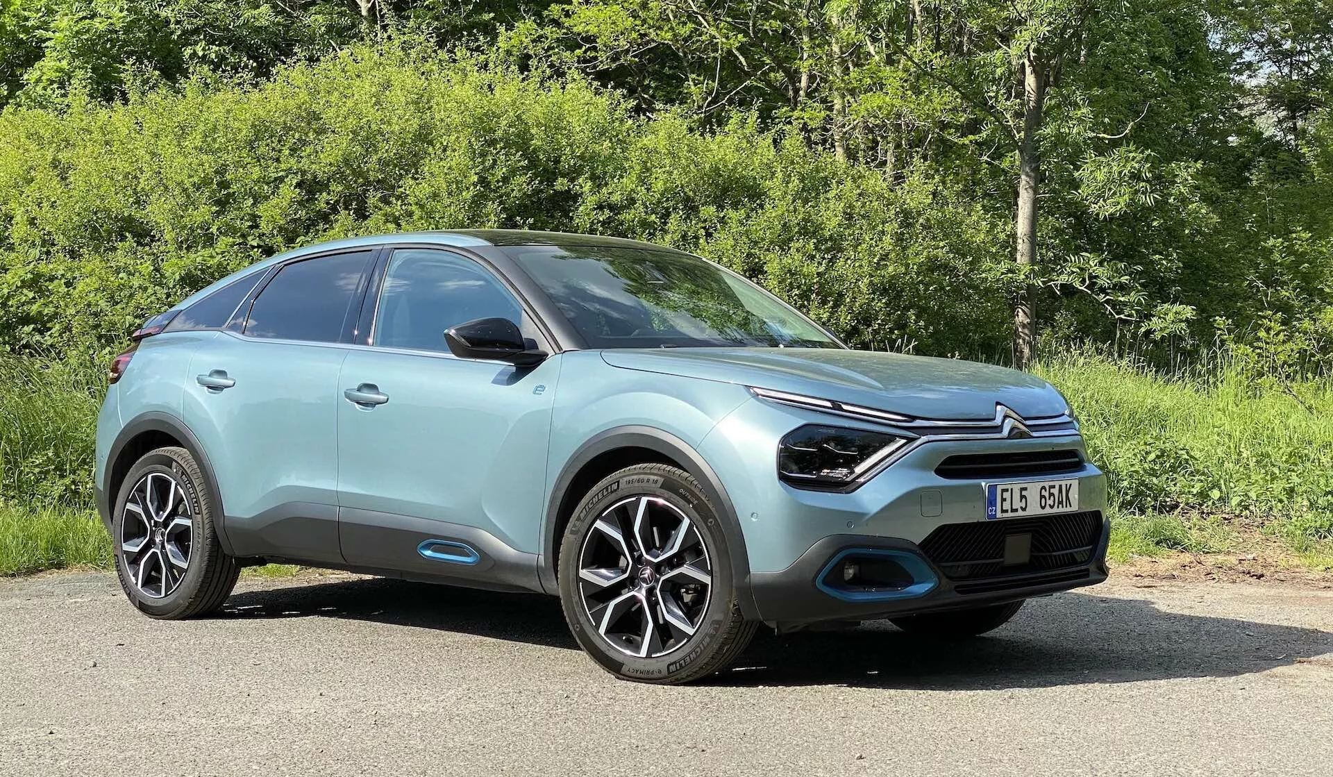 Pohled z nízkého úhlu na přední a boční část Citroenu C4 roku 2020 (e-C4) zaparkovaného venku, v přírodním prostředí s rostlinami v pozadí. Ukazuje elegantní, aerodynamický design, výraznou přední masku s centrálně umístěným emblémem, úzké LED světlomety, integrované mlhovky v černých vložkách, vícepaprsková litá kola, lesklou mátovou nebo světle tyrkysovou barvu karoserie, hladké tvarované linie na kapotě a bočních panelech, zrcátka s integrovanými blinkry, mírně se svažující střechu naznačující karoserii typu crossover SUV, černé obložení kolem podběhů kol a podél spodní části karoserie a část čelního skla odrážející jasnou oblohu za slunečného dne.