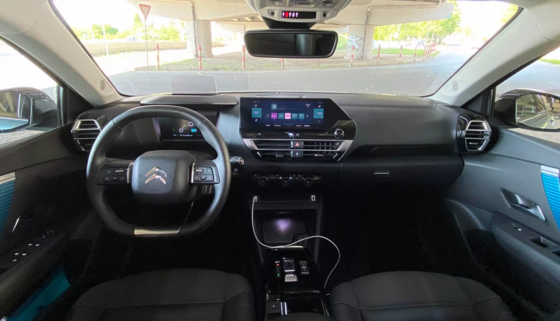 Interiér modelu Citroen C4 2020 (e-C4) pohledem ze zadního sedadla, zaměřený na palubní desku s digitálním přístrojovým klastrem, moderním volantem s vestavěnými ovládacími prvky a prominentní informační obrazovkou ve středové konzoli. Konzole obsahuje tlačítka klimatizace, řadicí páku a další ovládací prvky, které naznačují funkce jako GPS navigace, multimediální systém a manuální nebo automatickou klimatizaci. Přední sedadla jsou čalouněná s opěrkami hlavy a dveřní panely mají prémiové akcenty.