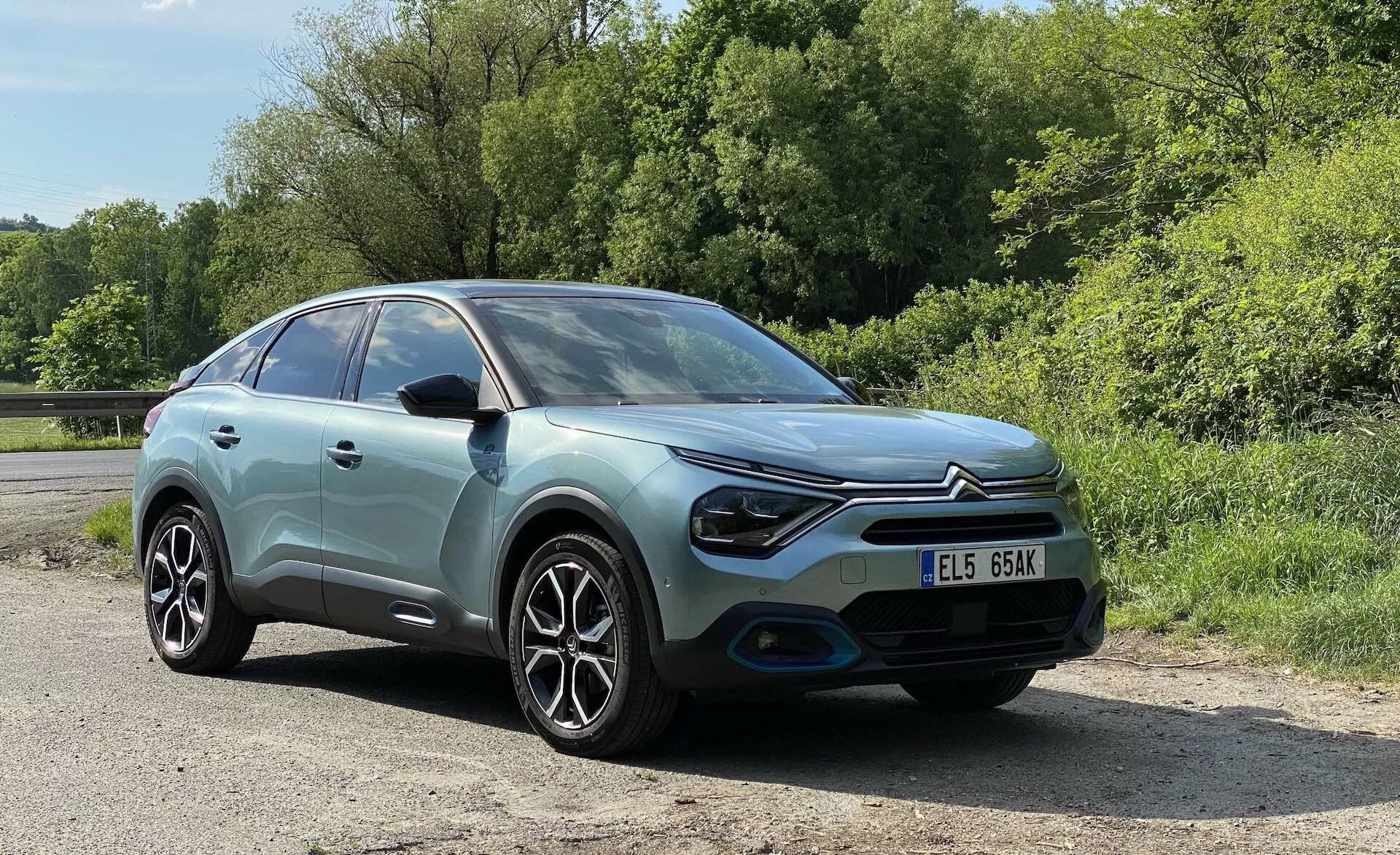 Obrázek Citroenu C4 2020 (e-C4) zachycený z tříčtvrtečního čelního pohledu, zvýrazňující pastelově modré tělo s kontrastní černou střechou, moderními LED světlomety, tvarovaný přední nárazník, hliníkové kola a složitý vzorek paprsků. Obrázek, pořízený z mírného úhlu dolů, poskytuje podrobné pohledy na levé světlomety, přední nárazník, přední kolo, čelní sklo a okno řidiče, s registrační značkou zřetelně viditelnou. Auto je umístěno na pozadí zeleně v přírodním nebo parkovém prostředí, za slunečného denního světla.