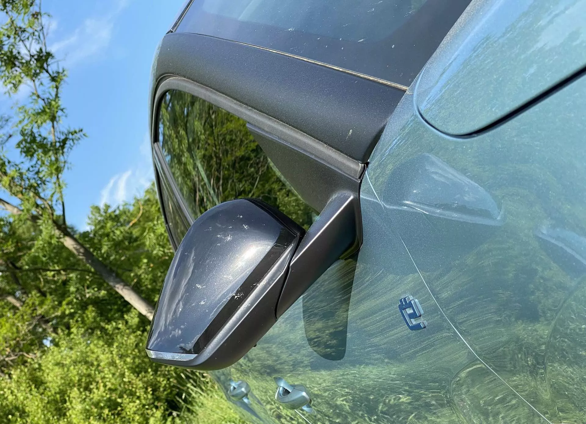 Detailní pohled na Citroen C4 2020 (e-C4) zřidičovy strany focený z nadhledu, zaměřený na moderní boční zpětné zrcátko s integrovanými směrovými ukazateli, kryt barevně sladěný s karoserií a v tónovaných oknech odráží venkovní zeleň. Světlá denní scéna zvýrazňuje metalický lak a elegantní moderní design vozu.