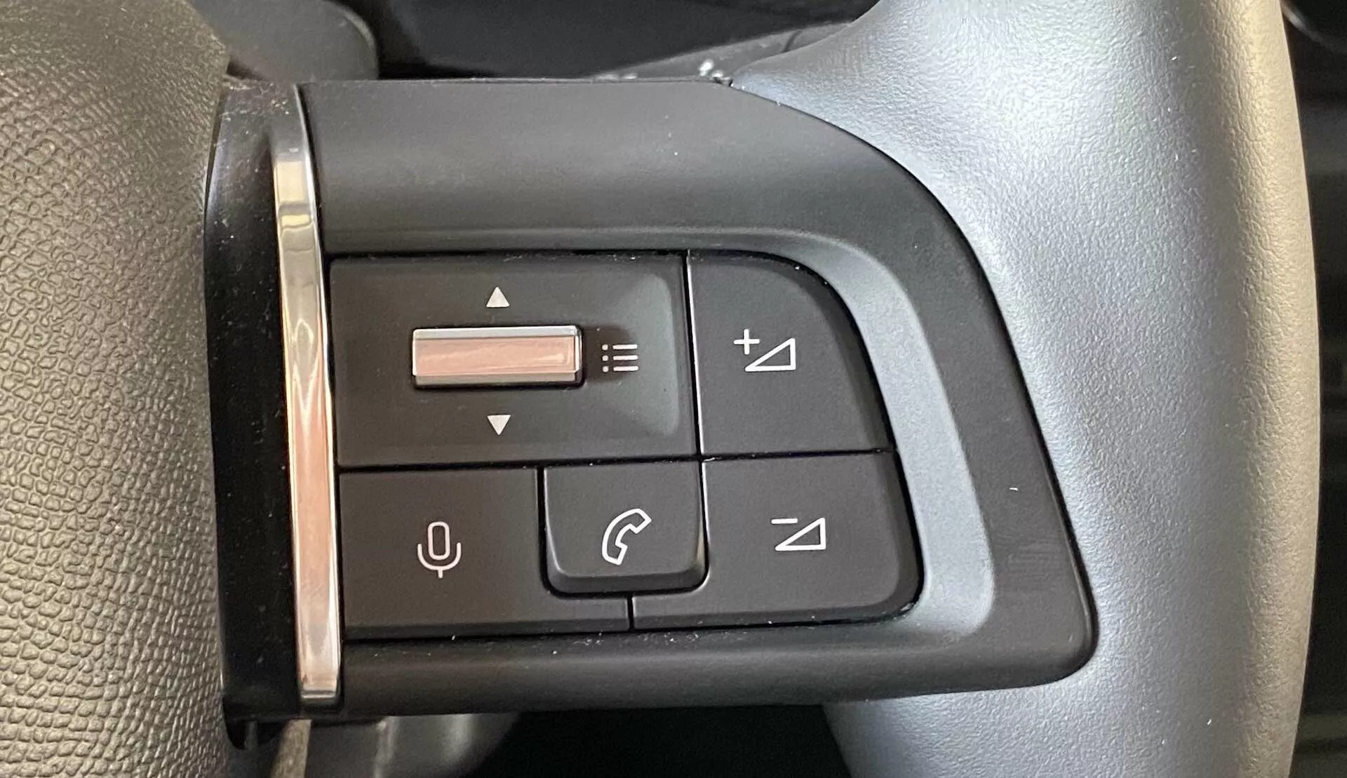 Detailní pohled na multifunkční ovládací panel na levé straně volantu v Citroënu C4 2020 (e-C4) z pohledu řidiče. Panel obsahuje tlačítka se symboly pro ovládání hlasitosti, přepínání skladeb, správu telefonních hovorů a malý joystick pro navigaci v menu. Černý panel a tlačítka jsou navrženy pro haptickou zpětnou vazbu a ladí s estetickým vzhledem volantu, který má texturovaný povrch a šev naznačující kvalitní materiál.