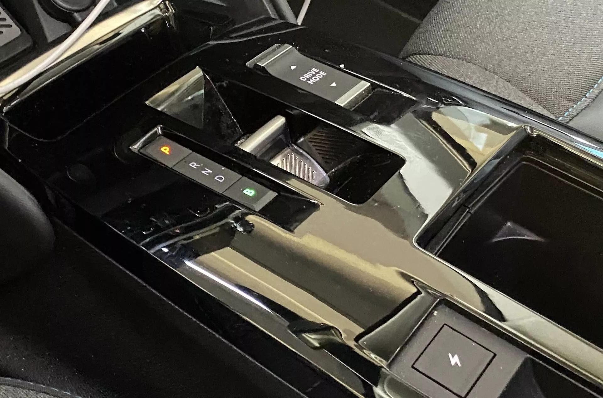 Detailní pohled na středovou konzolu uvnitř vozu Citroën C4 2020 (e-C4), zvýrazňující elektronický volič převodovky, tlačítka pro řízení jízdních režimů, držáky na nápoje, lesklý kryt úložného prostoru, elektronickou parkovací brzdu, lesklý černý materiál a částečně viditelné dotykové rozhraní, což naznačuje vysoce technologický, luxusní interiér vozu.