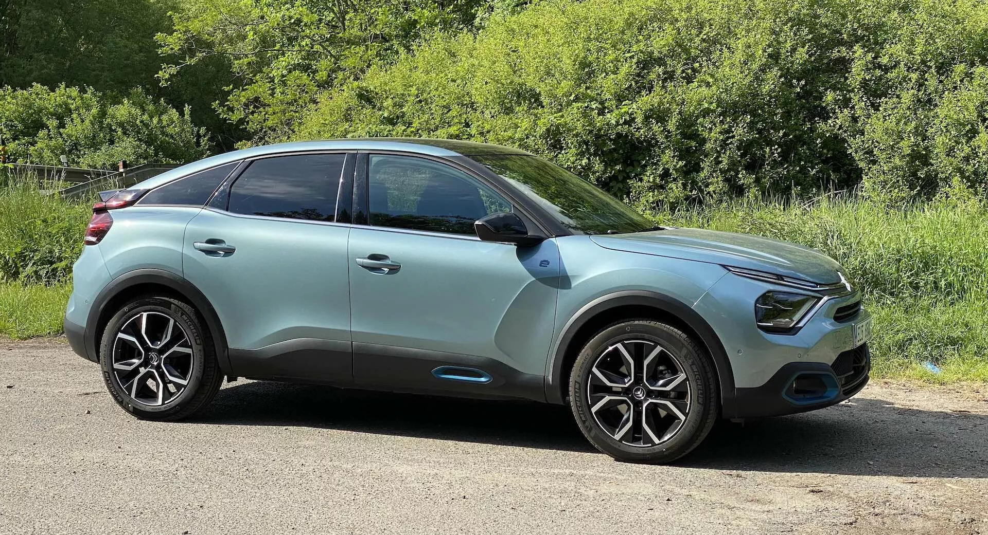 Boční profil Citroënu C4 2020 (e-C4) moderního crossover SUV pořízený z nižšího úhlu. Auto je zaparkováno na rovném kraji silnice s zelení v pozadí, což naznačuje venkovské nebo parkové prostředí. Elegantní kapota vede ke zakřivené přední mřížce, orámované LED denními světly a možnými mlhovkami. Dvoubarevné tělo v barvě světle tyrkysové s černým lemováním ukazuje pokročilé funkce jako integrované směrovky v bočních zrcátkách a senzory bezklíčového vstupu na barevně sladěných klikách. Velká, vícepaprsková slitá kola a vysoce výkonné pneumatiky zdůrazňují estetiku a výkon. Mírně klesající linie střechy, ztmavená okna a potenciální pokročilé systémy asistence řidiče ukazují zaměření na bezpečnost, pohodlí a aerodynamiku. Snímek pořízený za denního světla s autem umístěným v střední vzdálenosti zdůrazňuje design zevnějšku vozidla.
