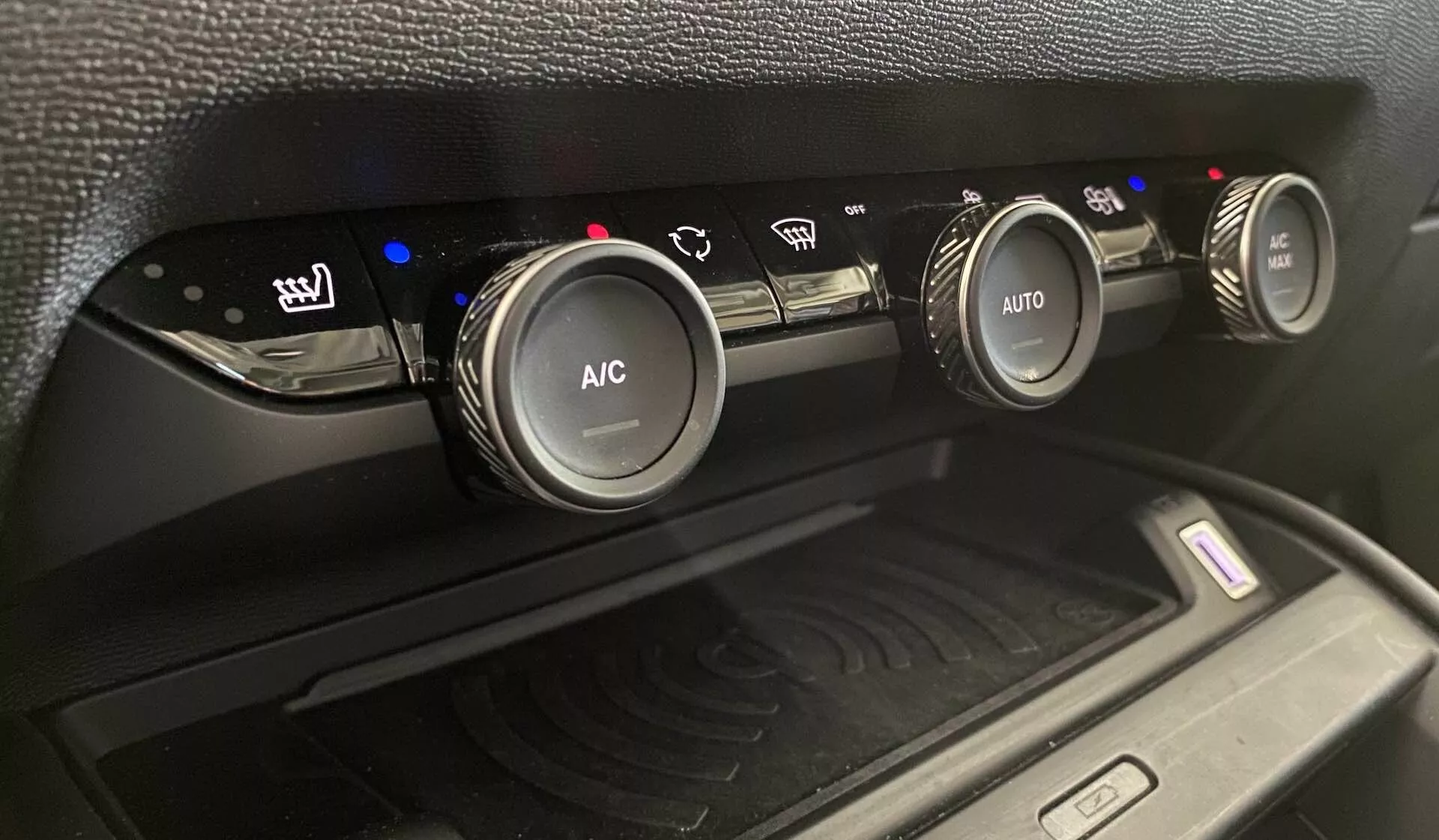 Detailní záběr klimatizačního systému vozu Citroen C4 z roku 2020 (e-C4), ukazující tři kruhové ovládací knoflíky na středové konzoli z mírně nakloněného úhlu nahoru. Levý knoflík ovládá vyhřívání sedadel s modrými indikátory úrovně tepla, středový knoflík je označen 'A/C' pro klimatizaci a pravý knoflík má 'AUTO' pro automatické nastavení klimatu. Modré a červené indikátory odrážejí nastavení teploty. Pod knoflíky je přihrádka s podložkou pro bezdrátové nabíjení s rádiovými vlnami, obklopená matně černým plastovým povrchem a texturou.