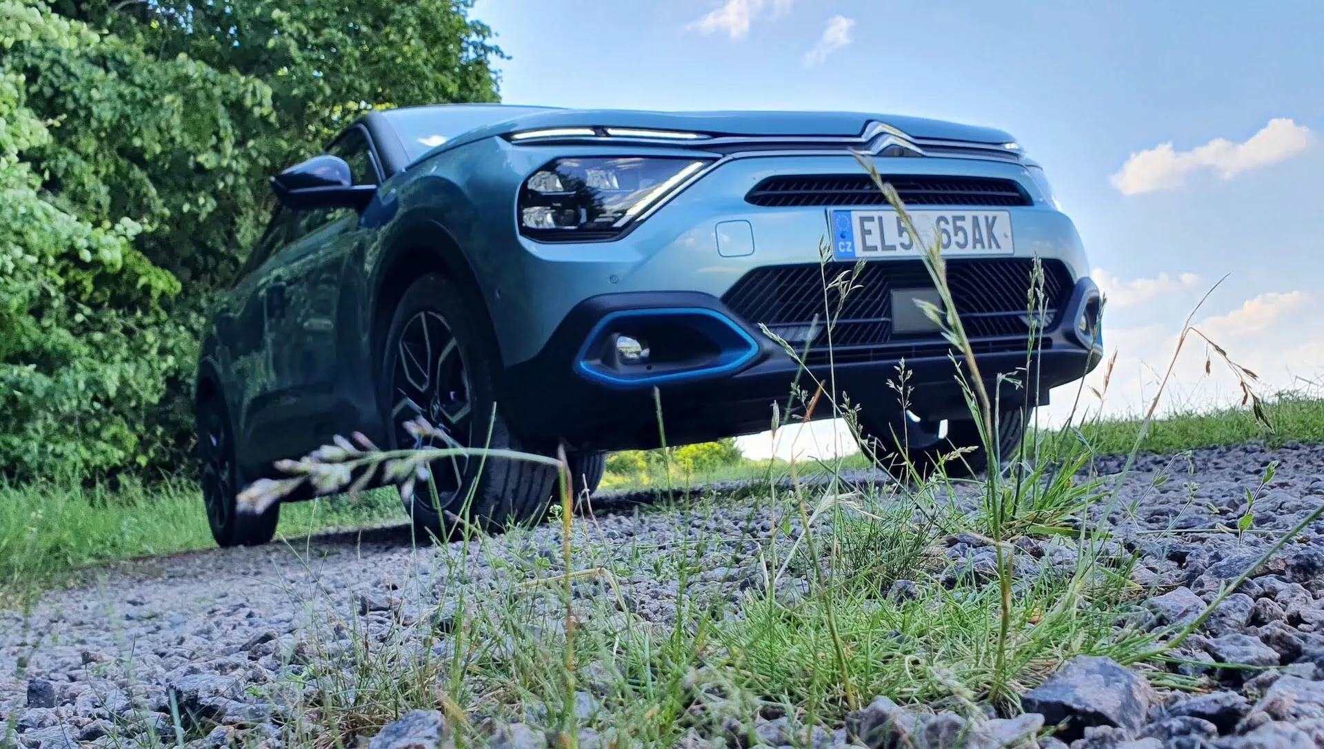Nízký čelní pohled na zaparkovaný Citroën C4 2020 (e-C4) na štěrkové cestě obklopené zelení, ukazující přední kapotu, mřížku chladiče, přední nárazník, světlomety a část předních kol. Obrázek zdůrazňuje robustní postoj vozidla a exteriérové designové prvky, jako je tvar světlometů, vzor mřížky, vzduchové vstupy a kontury kapoty. Sluneční světlo odrážející od lakování zvýrazňuje estetickou přitažlivost vozu. Potenciální pokročilé technologie osvětlení a parkovací senzory jsou rovněž součástí viditelných technologických prvků.
