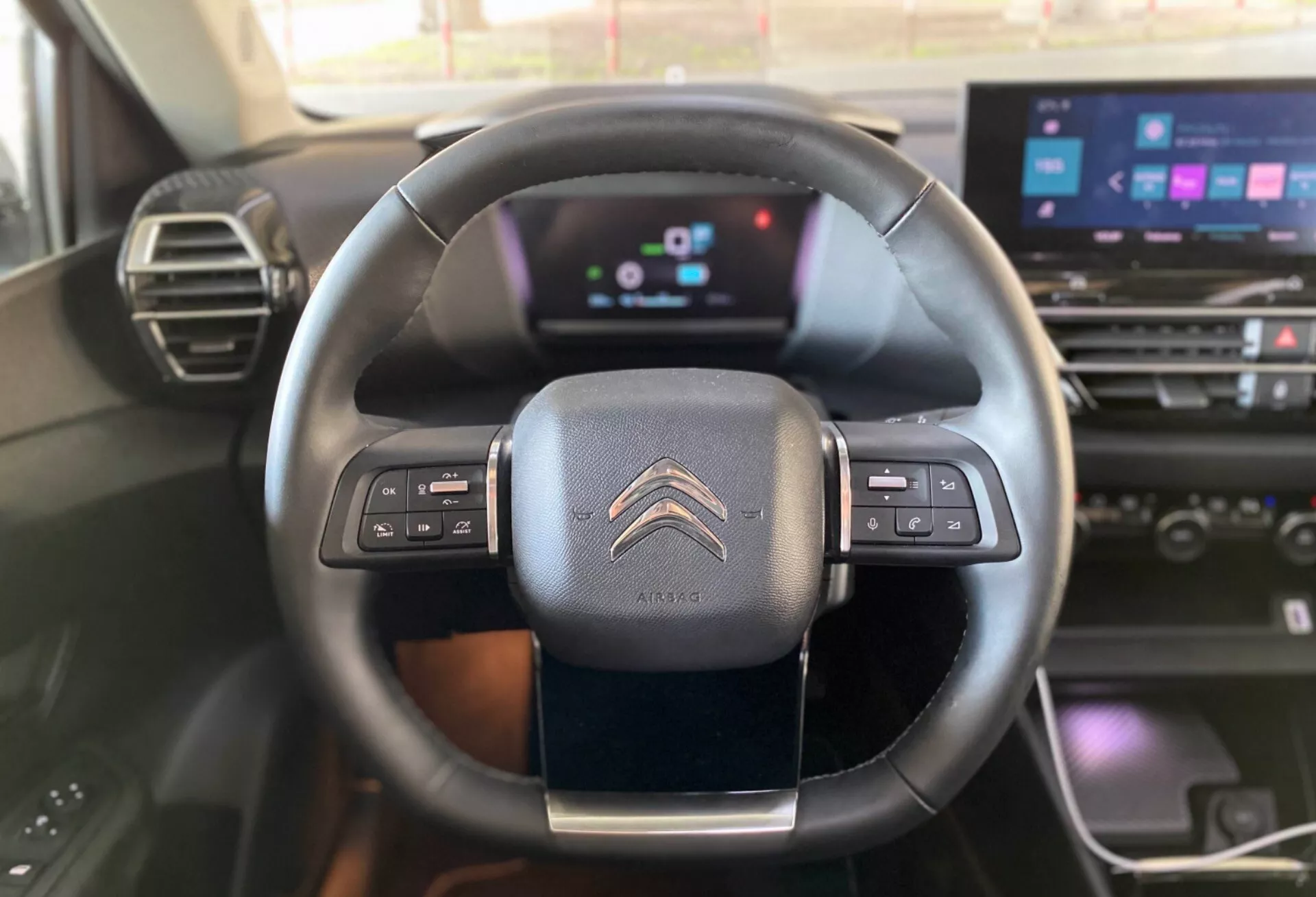 Pohled na interiér z perspektivy řidiče nebo spolujezdce modelu Citroën C4 2020 (e-C4), ukazující volant a moderní palubní desku. Tříramenný, kožený volant je vybaven ovládacími prvky pro audio, hlasové příkazy a tempomat. Digitální přístrojová deska nabízí barevné budíky a informační displeje, což značí pokročilou digitální technologii. Části infotainmentového dotykového displeje, tlačítka klimatizace a dřevěný akcent na palubní desce jsou viditelné, což zdůrazňuje technologicky vyspělý a esteticky příjemný interiér vozu.