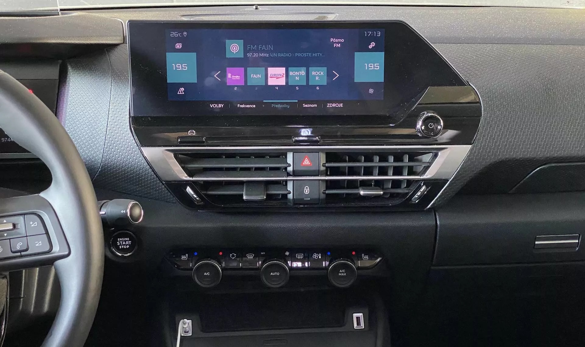 Pohled do interiéru Citroen e-C4 2020 zaměřený na palubní desku z centrální pozice. Obrázek ukazuje moderní dotykový infotainment displej s možnostmi navigace, médií, připojení telefonu a nastavení vozidla. Níže se nacházejí horizontální větrací otvory s tlačítky pro klimatizaci a další funkce a dvojzónová klimatizace s otočnými ovladači teploty a LCD displejem. Část volantu s montovanými ovladači a start-stop tlačítkem je viditelná, zdůrazňující moderní technologii ve voze a ergonomický design.