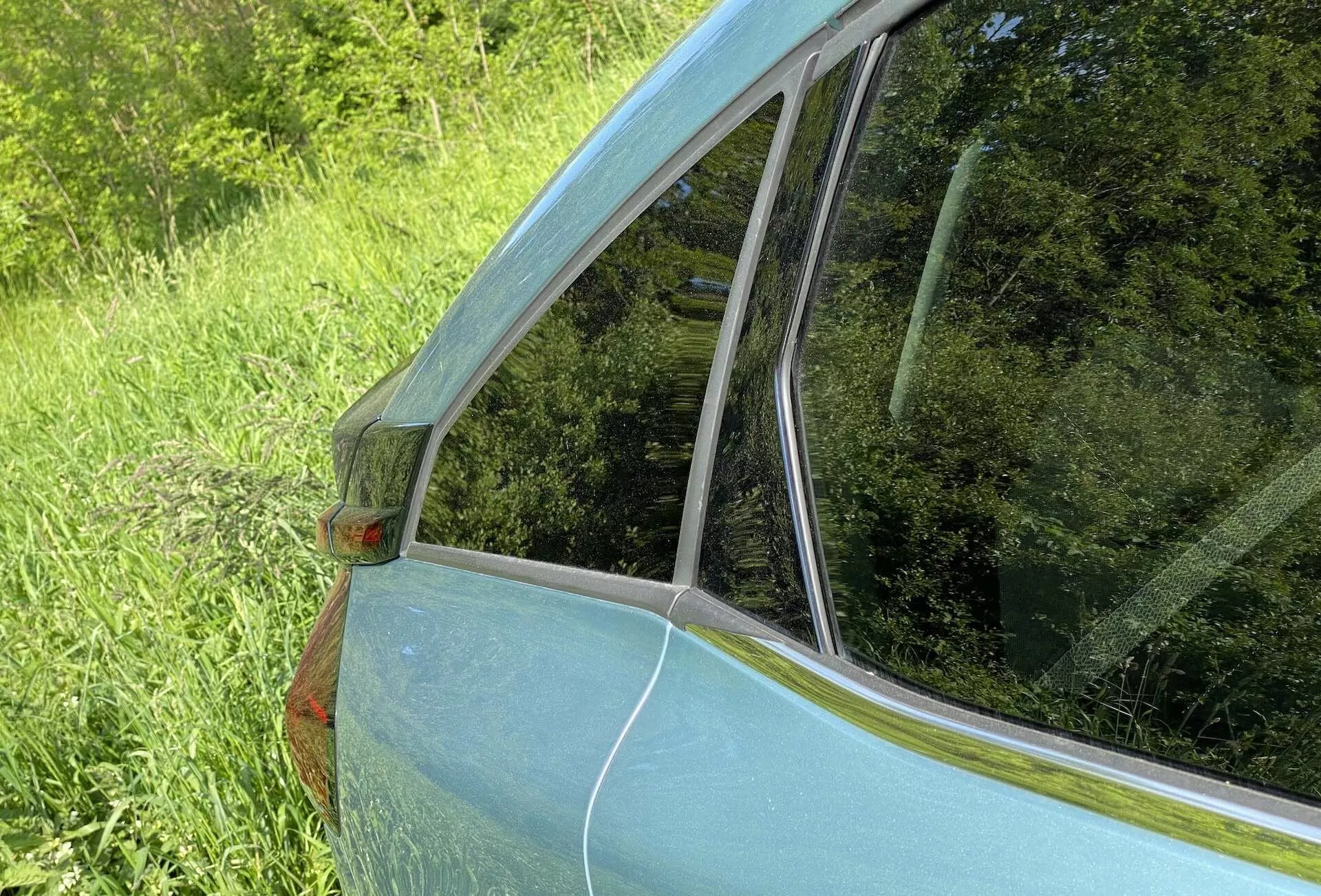 Detailní pohled na zadní boční panel a okno Citroenu C4 2020 (e-C4). Snímek pořízený ze šikmého úhlu ze zadní strany vozu zdůrazňuje přechod od zadního okna pasažéra k zadnímu sklu. Viditelný je kovový světle modrý lak, oranžové boční odrazové světlo na C-sloupku, chromové lemování a odrazy na skle, zdůrazňující design a stav vozu.