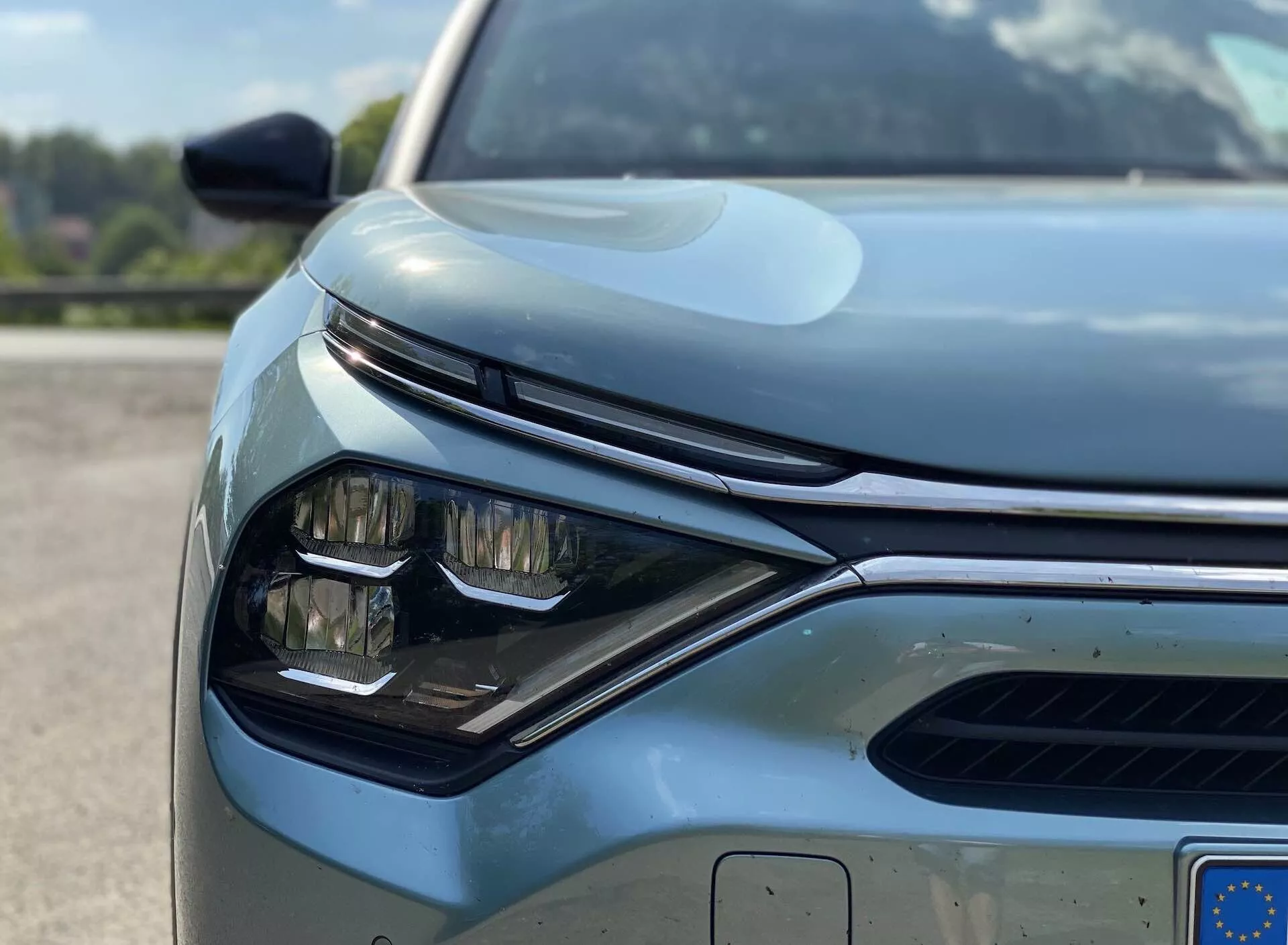 Zblízka, z nízkého úhlu pohledu na přední levý čtvrt vozidla, konkrétně Citroen C4 2020 (e-C4). Zaměřeno na elegantní, hranatý design světlometu s viditelnými LED prvky, část mřížky s horizontálními lamelami pravděpodobně s aktivním systémem řízení proudu vzduchu, a parkovací snímače na předním nárazníku. Na obrázku jsou zachyceny části bočního zrcátka vybaveného směrovými světly nebo snímači pro monitorování mrtvého úhlu. Kapota má hladké obrysy s vyvýšenou centrální částí, což dodává vozidlu jemně sportovní vzhled. Dobře udržovaný, reflexní povrch barvy naznačuje vysoce kvalitní materiál nebo účinný ochranný nátěr. Částečně viditelná evropská registrační značka zdůrazňuje moderní automobilovou technologii a design.