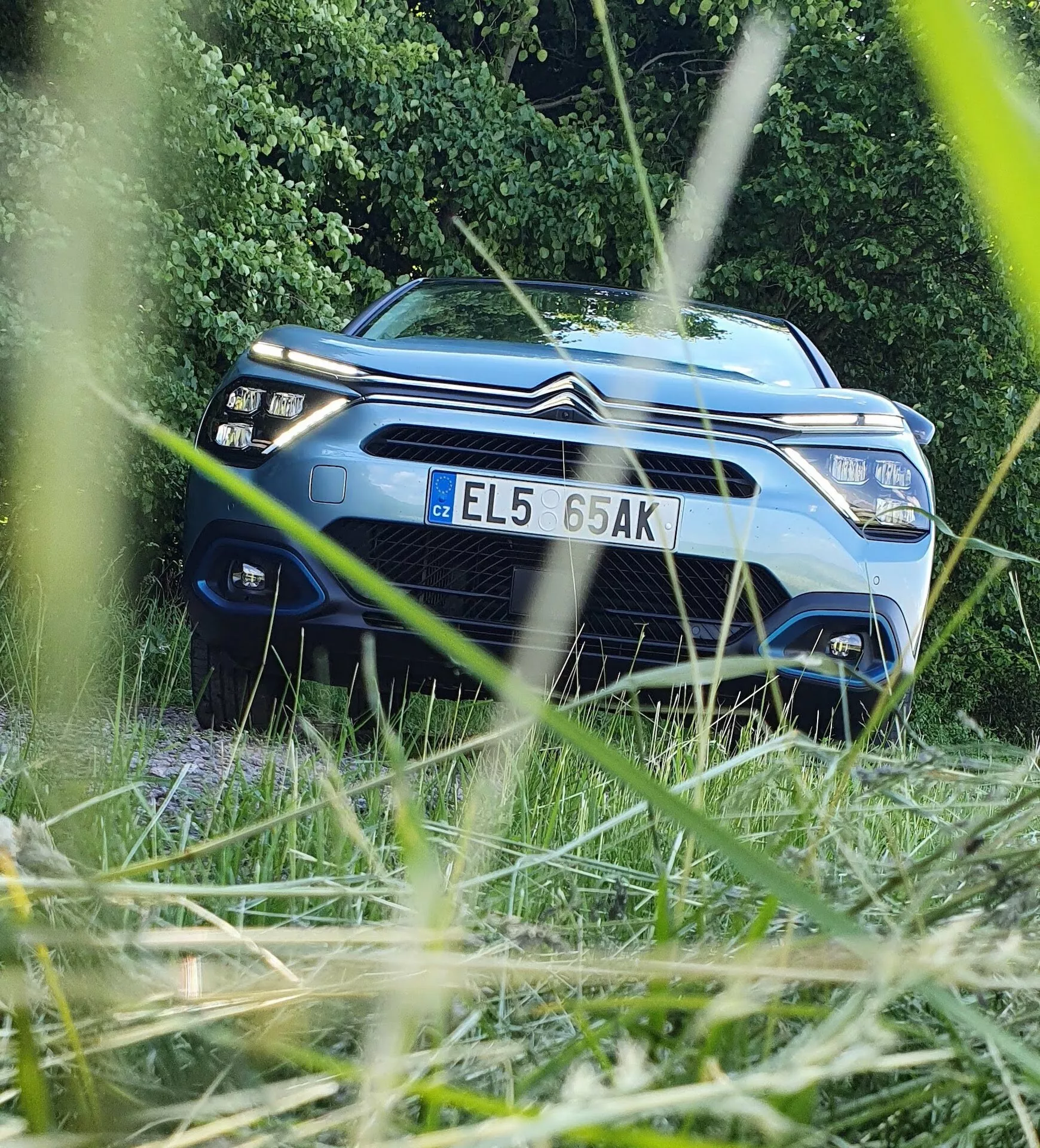 Modrý Citroën C4 2020 (e-C4) s výrazným duálním logem ve tvaru dvou šípů na přední mřížce, zachycený z nízkého předního úhlu, zasazený do rámu zelených listů v pozadí. Obrázek zdůrazňuje moderní světlomety vozu, elegantní přední fasádu a zvýšenou centrální část kapoty. Mělká hloubka ostrosti fotografie přivádí vůz do ostrého zaostření, zatímco nepatrně rozostřuje bezprostřední okolí, čímž zdůrazňuje designové a technologické prvky vozu.