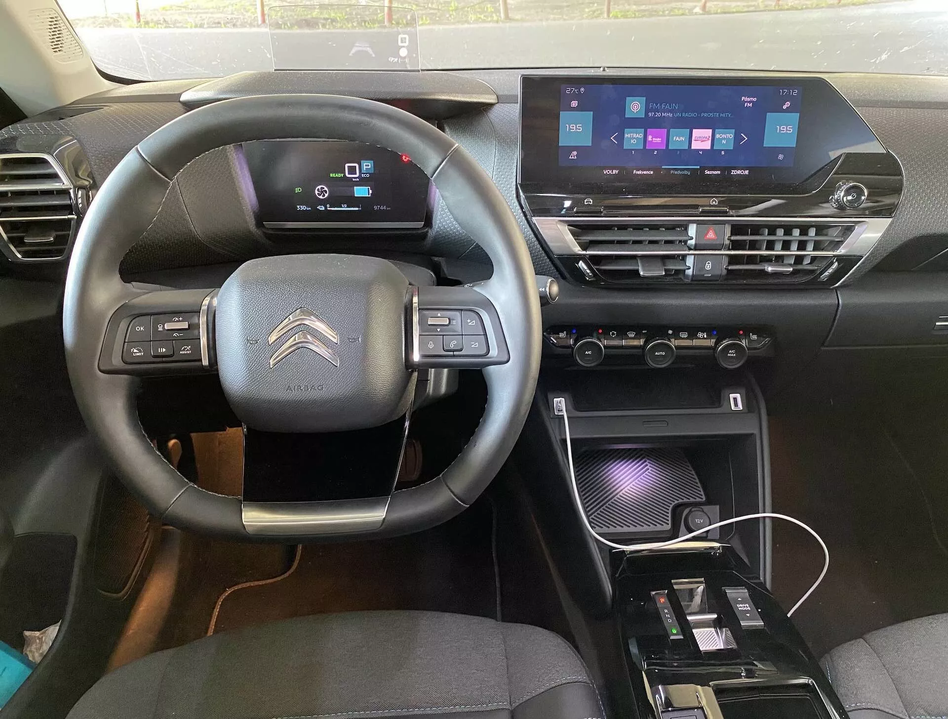 Pohled na interiér Citroënu C4 2020 (e-C4) z pozice řidiče, zobrazující tříramenný volant s ovládacími prvky, digitální přístrojový panel a moderní středovou konzoli s dotykovou informační jednotkou, ovladači klimatizace a nabíjecí oblastí.