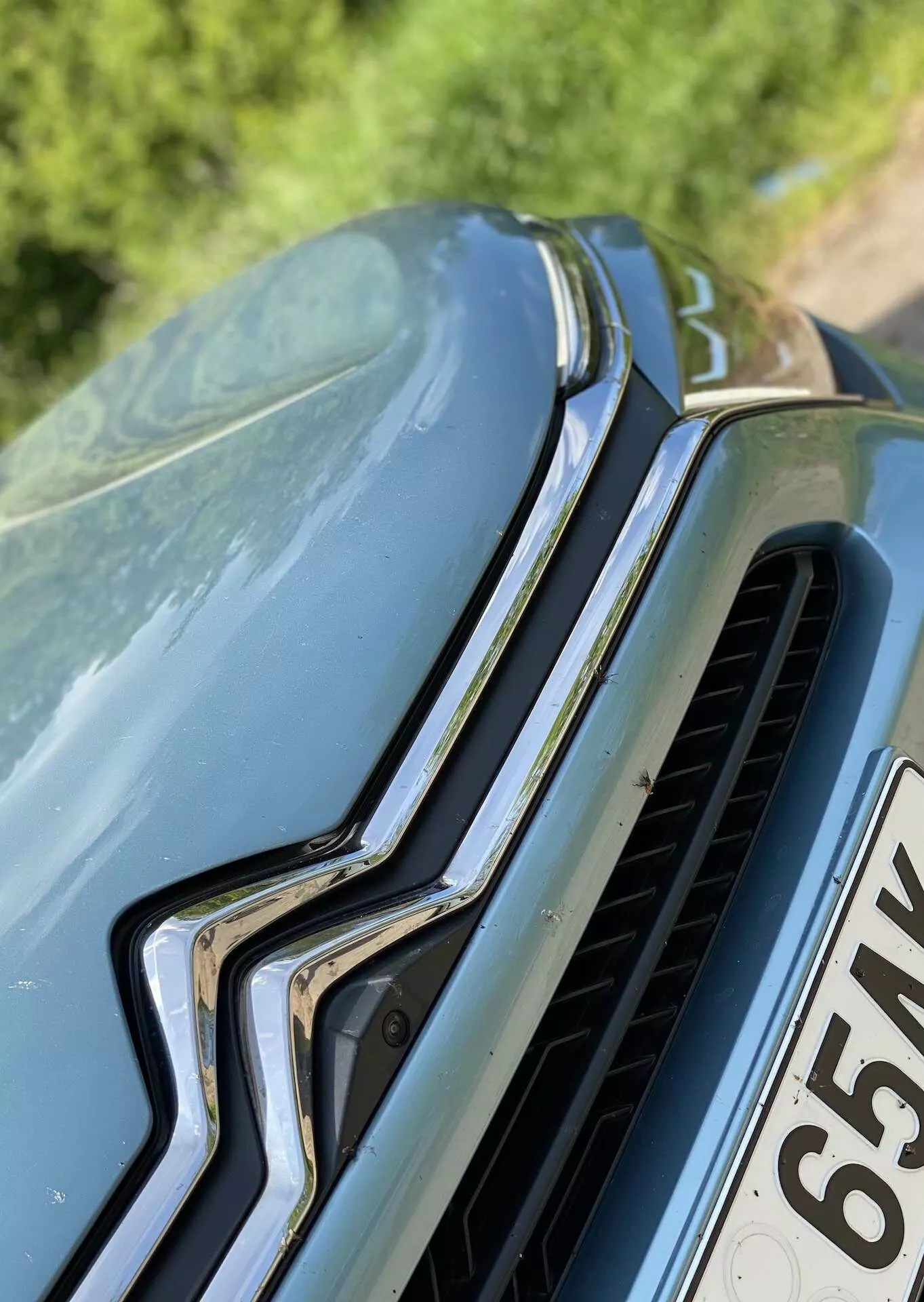 Detailní pohled na auto z předního a mírně výše položeného úhlu, zvýrazňující přední mřížku s vertikálně orientovaným chromovým kusem a horizontálními lamelami, výraznou značku na vrcholu a tvarovanou kapotu s vyvýšenými konturami. Viditelná špína nebo pyl na kapotě naznačují venkovní prostředí.