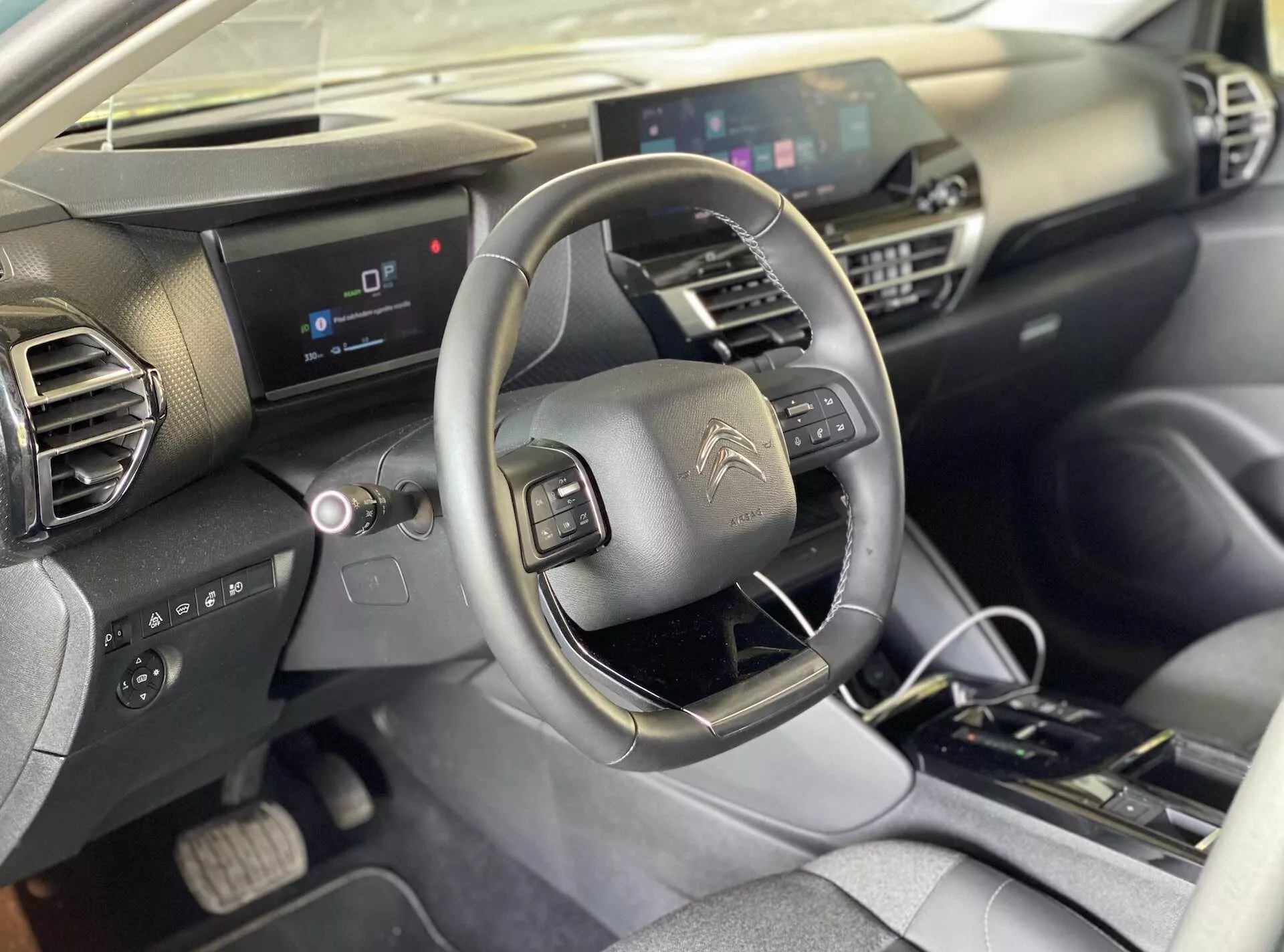 Pohled na interiér Citroënu e-C4 2020 z perspektivy předního spolujezdce, zachycující řidičovo sedadlo, středovou konzolu, volant s integrovanými ovládacími tlačítky, digitální přístrojový panel a centrální infotainment obrazovku. Palubní deska má geometrický design s tmavými a světlými materiály. Středová konzole obsahuje fyzická ovládání klimatizace a automatickou řadicí páku.
