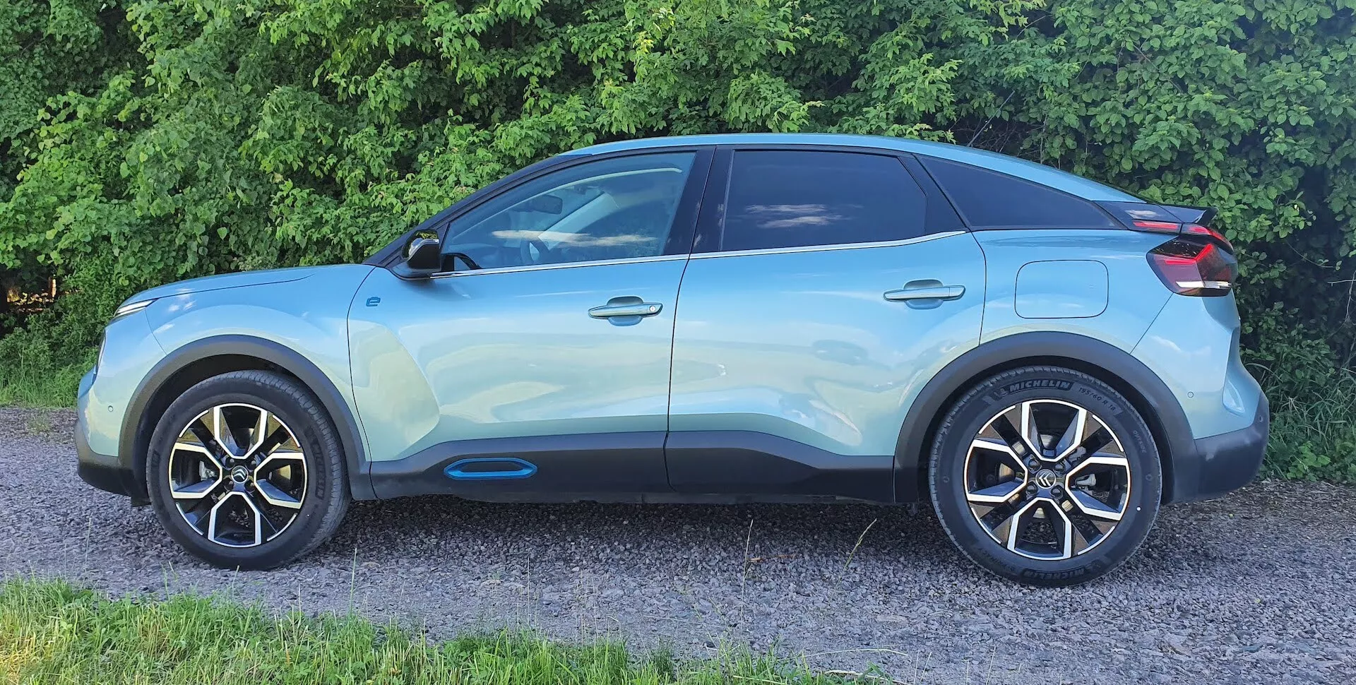 Boční profilový pohled na moderní crossover SUV Citroen C4 (e-C4) z roku 2020. Světle modré vozidlo je fotografováno z bočního-předního úhlu, zdůrazňující detaily jako lesklý lak, litá kola, boční zrcátka v barvě karoserie, výrazné charakteristické linie, černé lemování oken, tónovaná zadní okna, zapuštěné kliky dveří s bezklíčovým vstupem a svažující se střešní linii s možným střešním spoilerem a střešními nosiči. Na obrázku jsou zřetelně viditelné přední kolo a blatník, ale nejsou vidět přední maska, nárazník a světlomety.