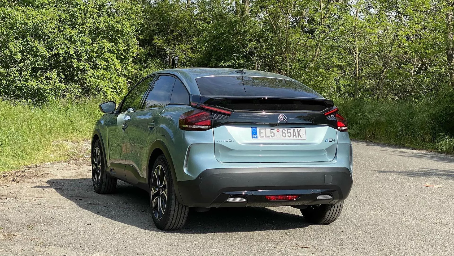 Snímek zadního úhlu zaparkovaného Citroenu C4 2020 (e-C4) na asfaltu s přírodními listy v pozadí, který ukazuje jeho výrazné LED zadní světla, značkování a model, zadní sklo s stěračem, dvoutónový nárazník s prvkem podobným difuzoru, integrované parkovací senzory, částečně viditelný výfuk, střešní anténu, vícepaprsková slitinová kola, metalickou povrchovou úpravu laku a evropskou registrační značku.