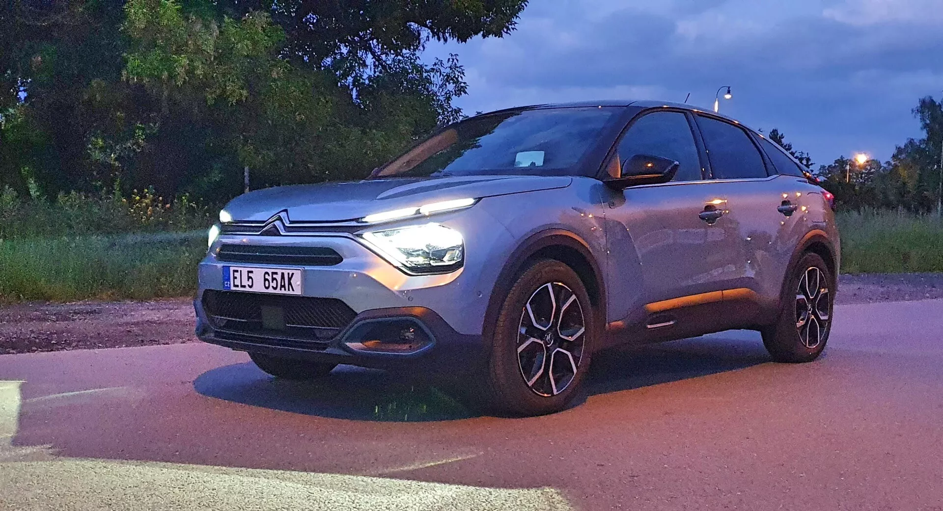 Citroen C4 2020 (e-C4) moderní crossover SUV zobrazený za soumraku s rozsvícenými hlavními světly a denními svítilnami. Obrázek zobrazuje čelní fasádu, mřížku s emblémem značky, kapotu, přední nárazník a boční profil řidiče včetně velkých vícepaprskových slitinových kol. Vozidlo, zabírající významnou část rámu, představuje LED světlomety, výraznou mřížku možná integrující senzory a kovový lak, což naznačuje vyšší úrovně výbavy nebo možnosti přizpůsobení.