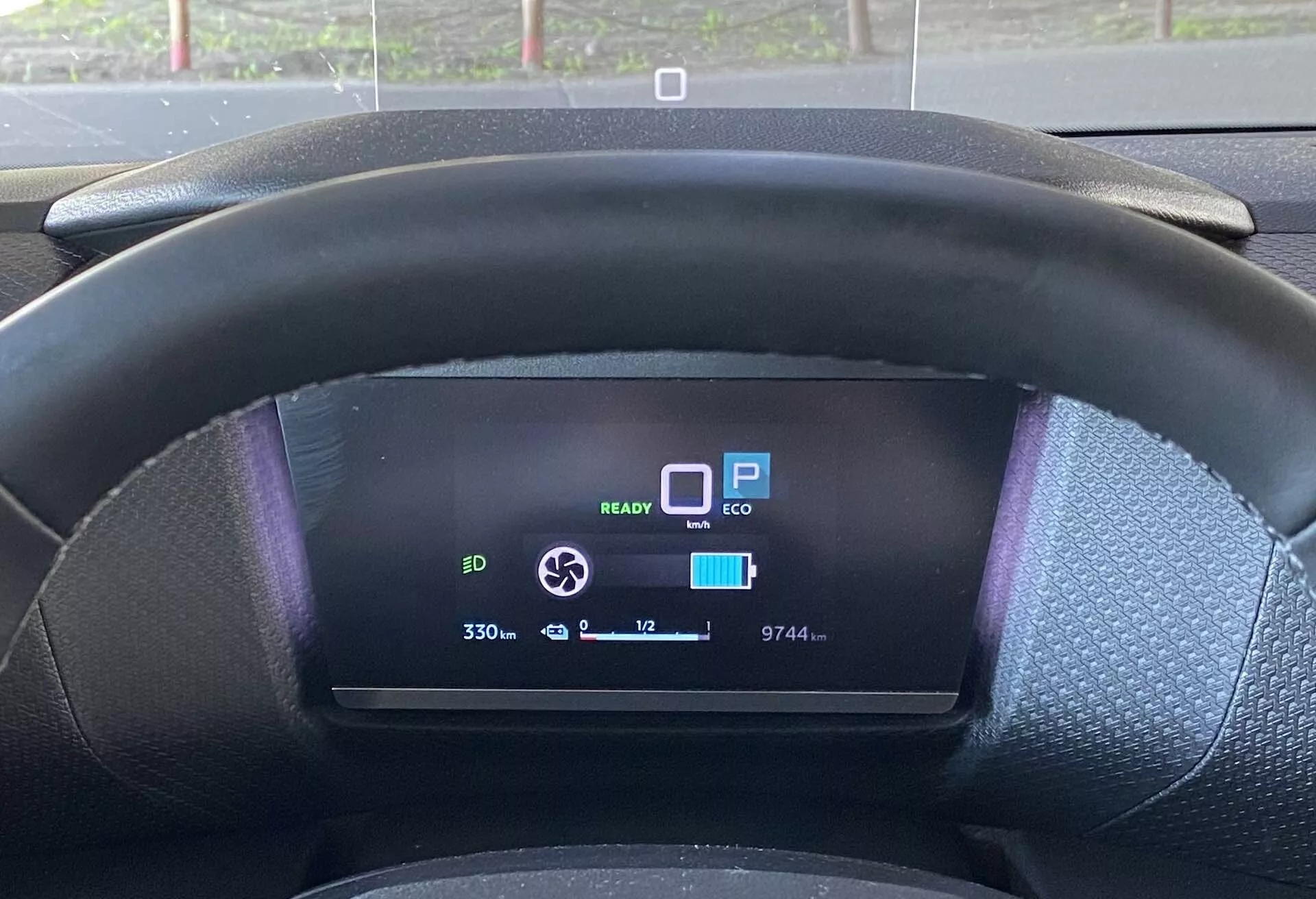 Detailní pohled z místa řidiče na Citroen C4 2020 (e-C4) zobrazující černý texturovaný volant a digitální přístrojový panel. Volant mírně zakrývá spodní část digitálního displeje, který zobrazuje několik funkcí: ikonu EV připravenosti 'READY,' 'P' pro parkování, režim 'ECO,' téměř plně nabitou ikonu baterie, ukazatel dojezdu '330,' odečet tachometru '9744,' venkovní teplotu '+12°C' a ikonu kruhové šipky, pravděpodobně pro nastavení rekuperačního brzdění.