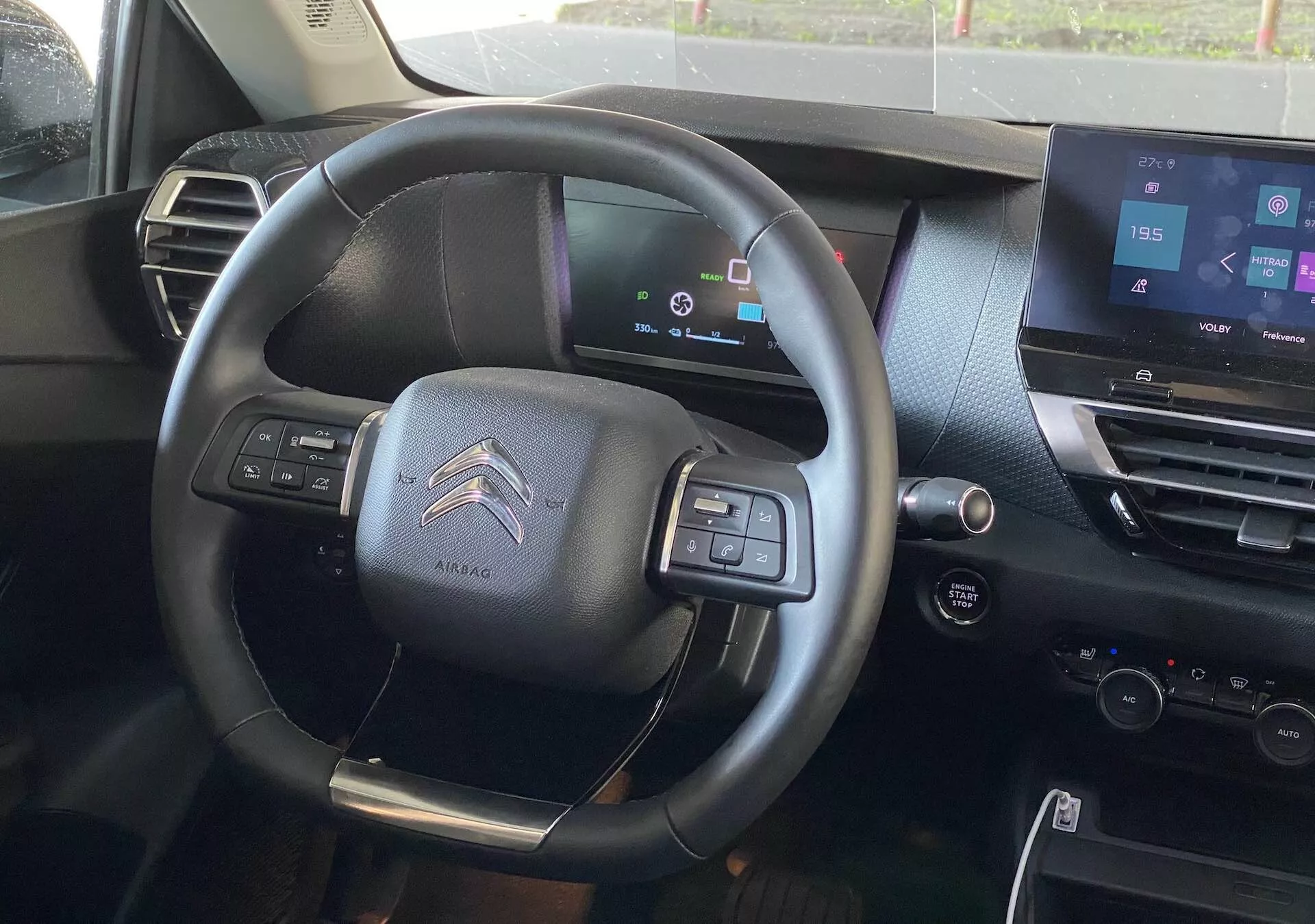 Pohled na interiér Citroënu C4 2020 (e-C4) z místa řidiče, se zaměřením na tříramenný multifunkční volant s airbagem, digitální přístrojovou desku zobrazující stav vozidla, dotykový infotainment systém na středovém panelu, tmavě laděný interiér, ovládací tlačítka pro audio a telefonní konektivitu, pádla řazení naznačující semiautomatickou převodovku a ovládací prvky na dveřích řidiče pro elektrická okna a nastavení zrcátek.