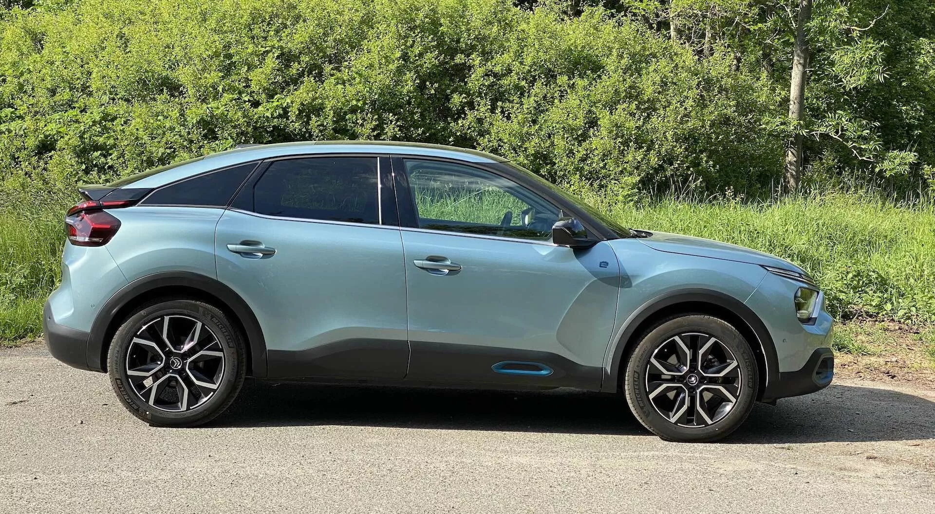 Boční profil Citroenu C4 (e-C4) z roku 2020, ukazující stranu řidiče z mírně úhlového pohledu zepředu. Obrázek zdůrazňuje moderní designové linie vozidla, litá kola s výrazným vícepaprskovým designem, boční zrcátka v barvě karoserie s integrovanými směrovkami a zadní okna s tónovaným sklem. Střecha se sklání směrem dozadu, dodávajíc vozu styl kupé na crossover/SUV těle. Auto má metalický lak s viditelnými obrysy a designovými liniemi, stojící na rovné ploše venku s pozadím zeleného listí, osvětlené přirozeným denním světlem.