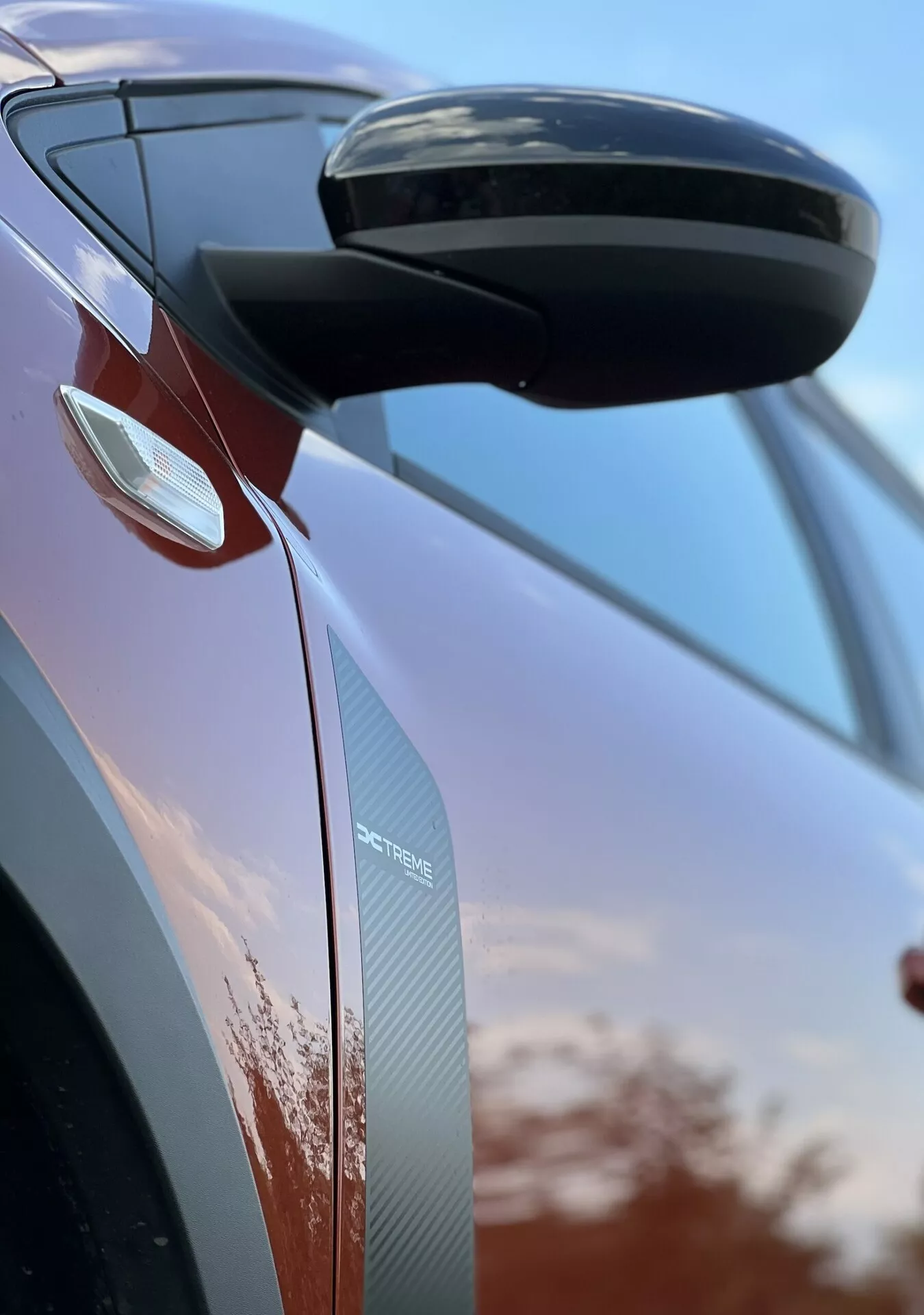 Detailní pohled na Dacia Jogger 2022 zaměřený na boční zpětné zrcátko a design dveří, který předvádí aerodynamické prvky, integrované směrové ukazatele a textury z uhlíkových vláken na pozadí oblohy