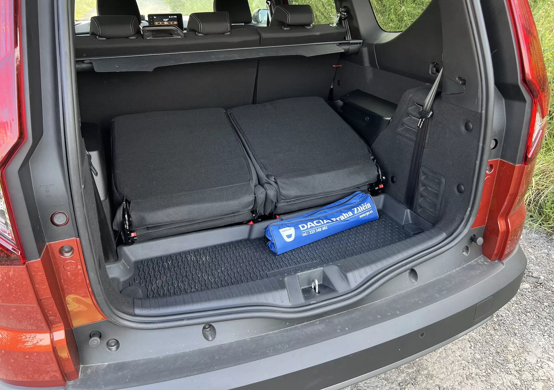 Detail kufru Dacia Jogger 2022 s otevřeným nákladovým prostorem, složenými zadními sedadly, textilními smyčkami pro zajištění předmětů a modrým válcem Decaux na kobercové podlaze