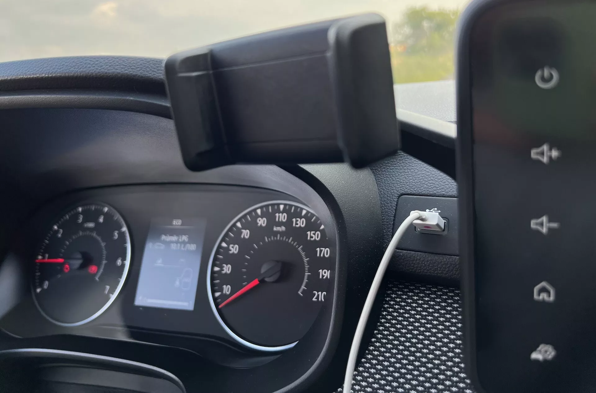Detailní interiérový pohled na palubní desku Dacia Jogger 2022 ukazující přístrojový panel, ovládání na volantu a část systému infotainment, naznačující funkce jako analogový tachometr, otáčkoměr, digitální displej pro 'Bez audia' a možnosti nabíjení smartphonu.