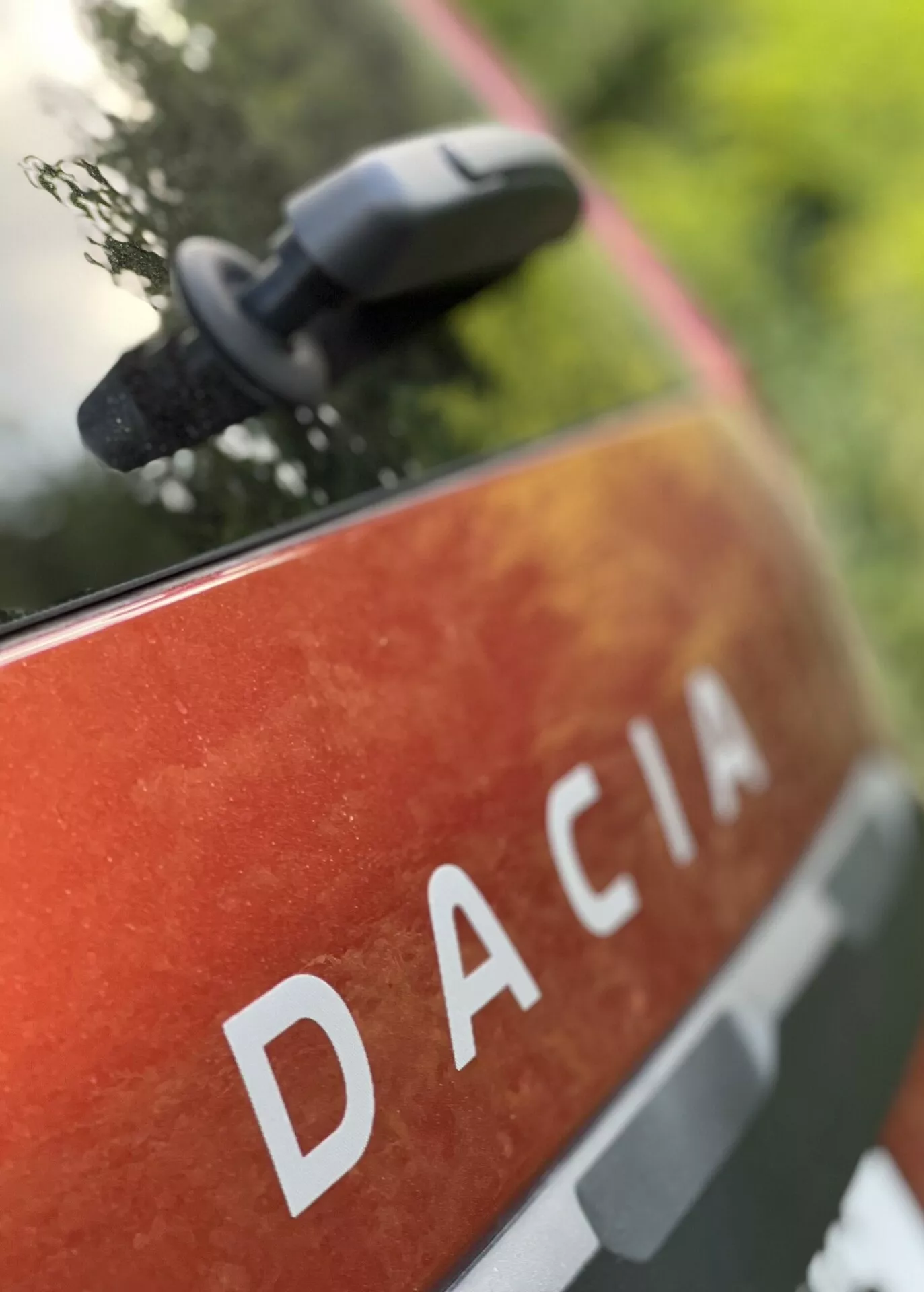 Detailní pohled na zadní panel Dacia Jogger 2022 s nápisem DACIA velkými písmeny bílé barvy na metalickém oranžovo-červeném laku s tečkami a nečistotami, naznačující venkovní použití a odolnost, umístěný v přírodním prostředí s rozmazaným bočním zrcátkem naznačujícím moderní funkce zrcátka.