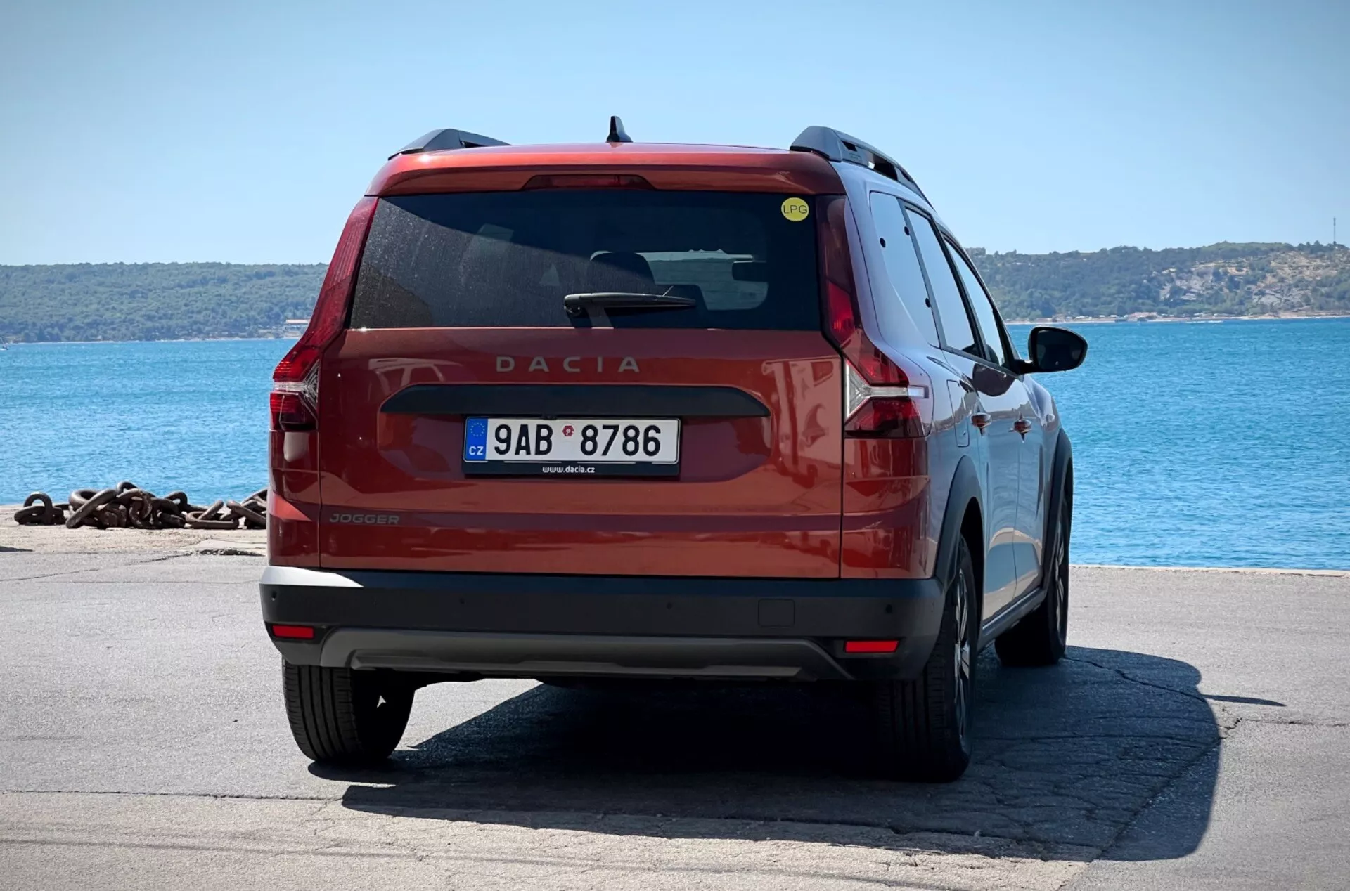 Červený Dacia Jogger 2022 fotografovaný z úhlu tři čtvrtě zezadu, předvádějící jeho zadní světla, stěrač zadního okna, označení modelu, nárazník a registrační značku na pozadí s vodní hladinou a vzdáleným pobřežím, zdůrazňující jeho design hatchbacku/SUV s potenciálními parkovacími senzory a pohledem na zadní dveře pro cestující, zadní blatník a část zadního kola.