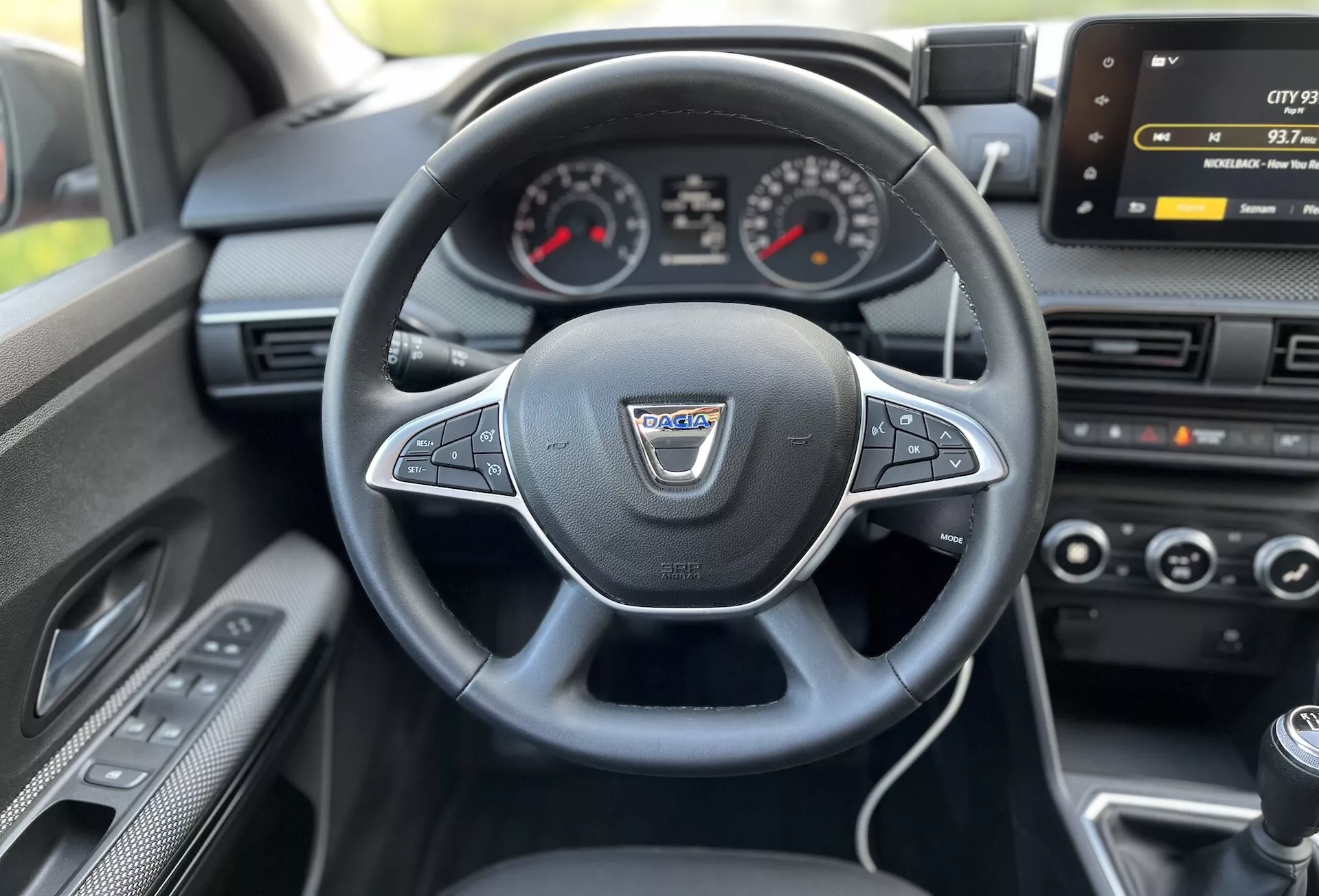 Interiérový pohled z místa řidiče na Dacii Jogger 2022 ukazující vícefunkční volant obalený kůží, analogový přístrojový panel s centrálním digitálním displejem, obrazovku infotainmentu, ovládání klimatizace a páku manuální převodovky