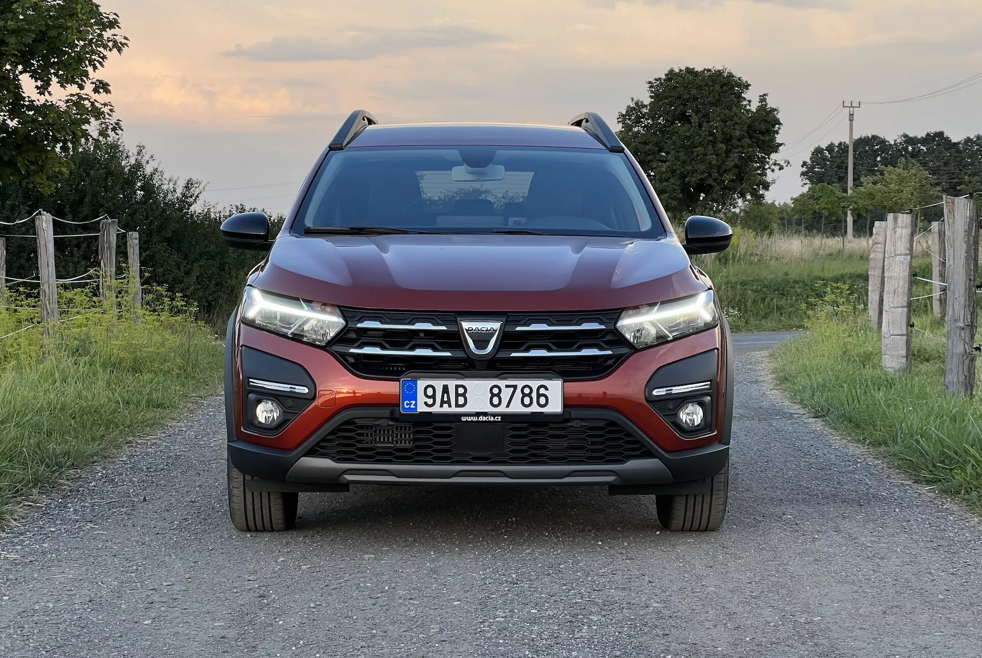 Dacia Jogger 2022 umístěný na úzké venkovské silnici, nabízí symetrický pohled zepředu, který zdůrazňuje moderní design vozu, včetně výrazné přední mřížky s velkým emblémem, štíhlých LED světlometů, konturovaných linií kapoty, předního nárazníku s mlhovkami a parkovacími senzory a zpětných zrcátek s integrovanými směrovými signály.