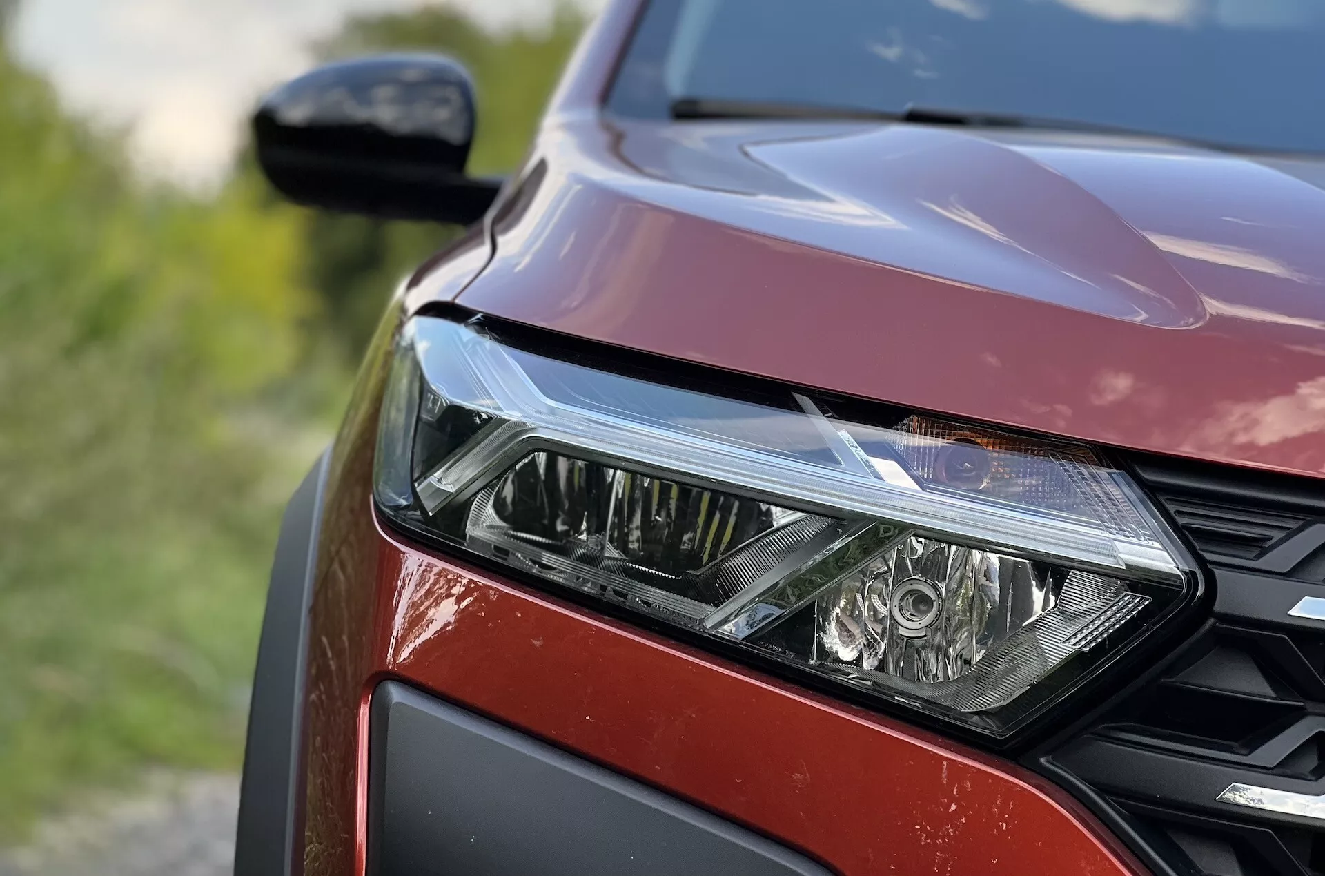 Detailní pohled na přední levou stranu Dacia Jogger 2022 zdůrazňující sestavu LED světlometů, vzor mřížky s medovými buňkami a metalický červený lak