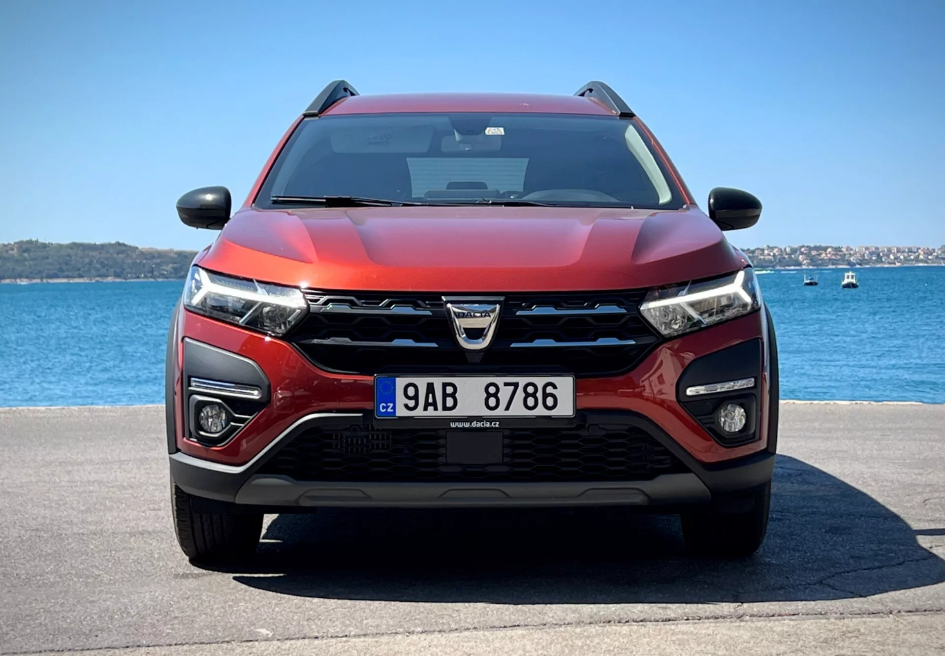 Dacia Jogger 2022 předvádí svůj moderní design crossoveru a pokročilé technologické funkce v klidném prostředí u vody, s výraznou maskou s logem, LED světlomety, mlhovými světly, potenciálními senzory ADAS, sacími otvory pro chlazení a radar, dvoubarevným červeno-černým barevným schématem, aerodynamickou kapotou, vybavená stěrači naznačujícími možnou technologii čištění deštěm, zpětnými zrcátky s integrovanými směrovými signály možná obsahujícími technologii monitorování mrtvého úhlu, pod jasným nebem.