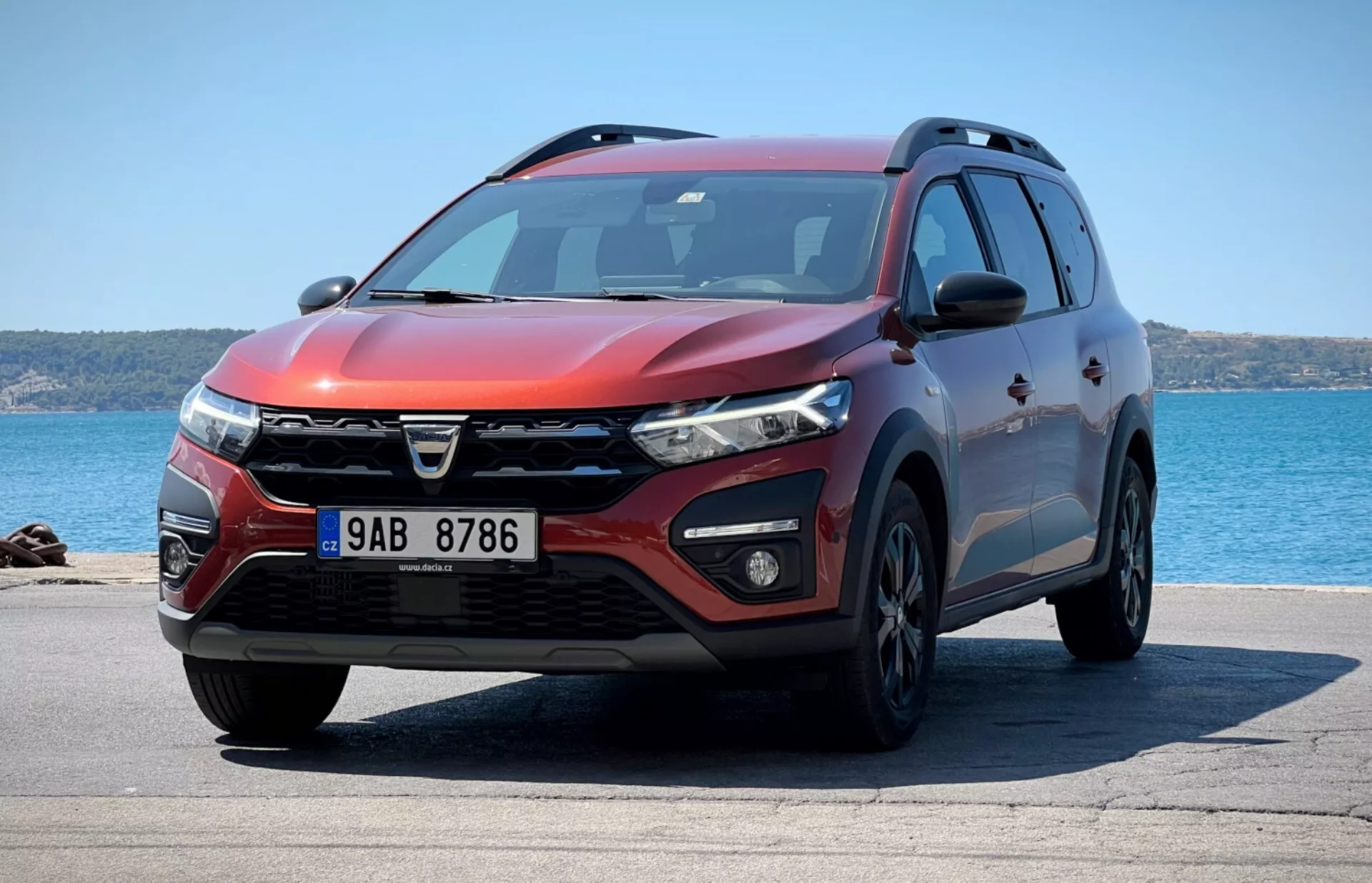 Čtvrtletní přední pohled na SUV Dacia Jogger 2022 zaparkované s výhledem na oceán, prezentující odlišný design mřížky, LED světlomety, mlhová světla, aerodynamický přední nárazník s vstupy vzduchu a parkovacími senzory, svalnatý kontur kapoty, slitinová kola, integrované směrové světla na bočních zrcátkách, boční profil s ochranným obložením, střešní nosiče, tónovaná okna a metalický červený exteriér pod jasným slunečním světlem ukazující designový profil, technologické funkce a užitkovost.