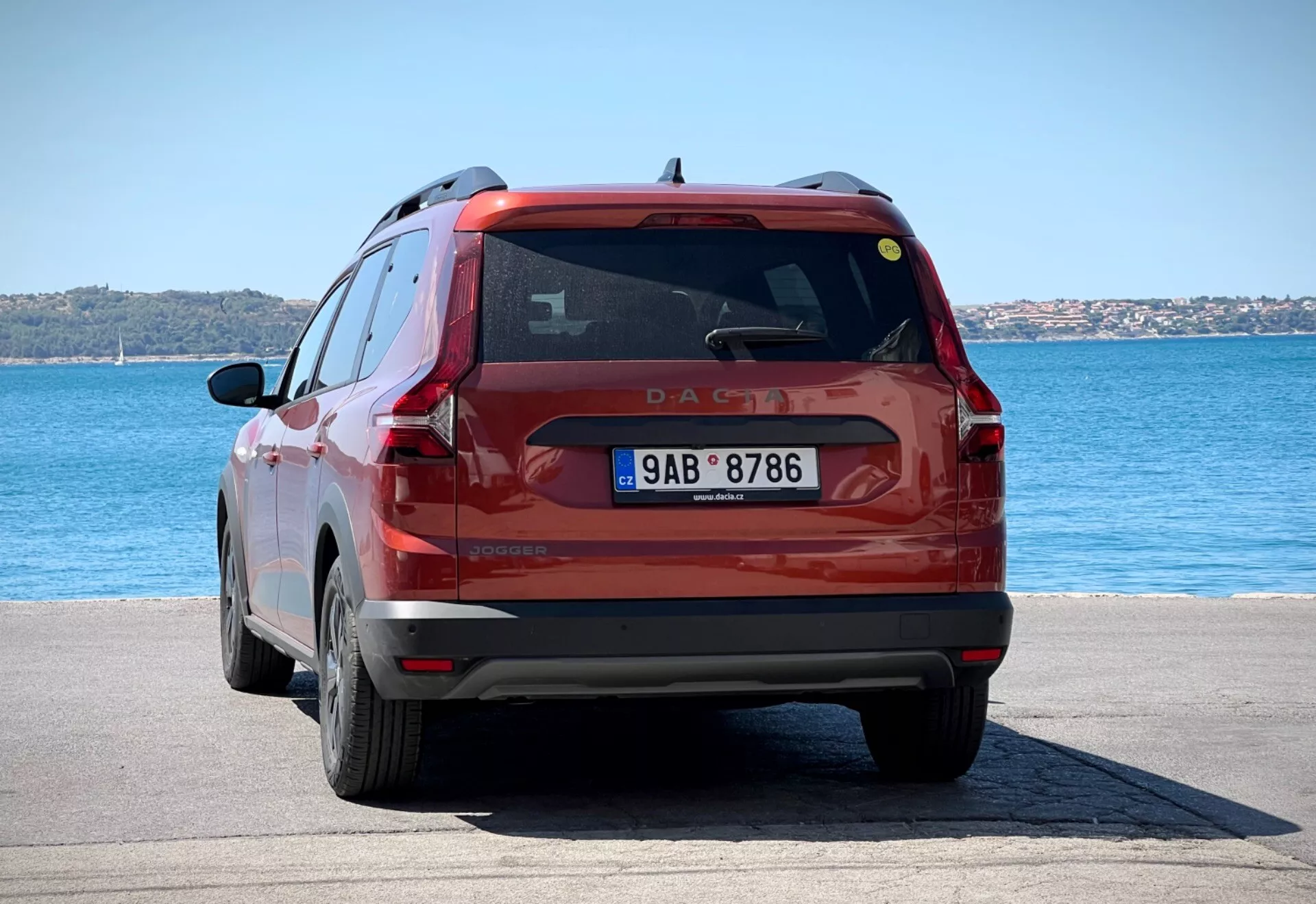Zadní pohled na červené kompaktní SUV Dacia Jogger 2022 zaparkované na dlážděném nábřeží pod jasným denním světlem, které zobrazuje celou zadní část vozidla včetně zadních světel, zadního okna a registrační značky na pozadí modré vody a jasné oblohy.