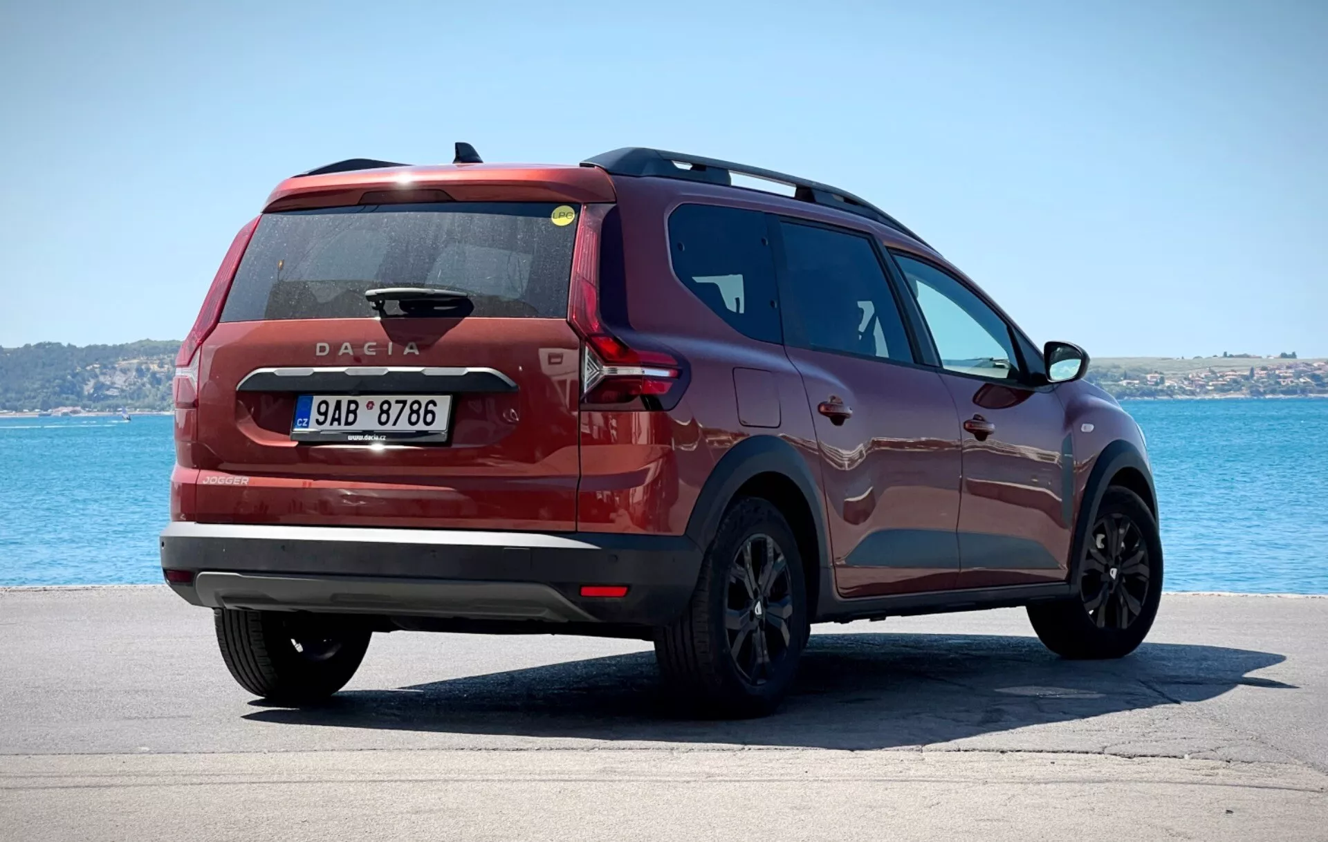 Dacia Jogger 2022 fotografována z trojčtvrtě zadního pohledu na dlažděném povrchu s tělesem vody a jasnou oblohou na pozadí, zobrazuje moderní zadní světla, centrálně umístěný znak Dacie a elegantní boční profil proti poklidnému pobřeží nebo jezernímu prostředí.